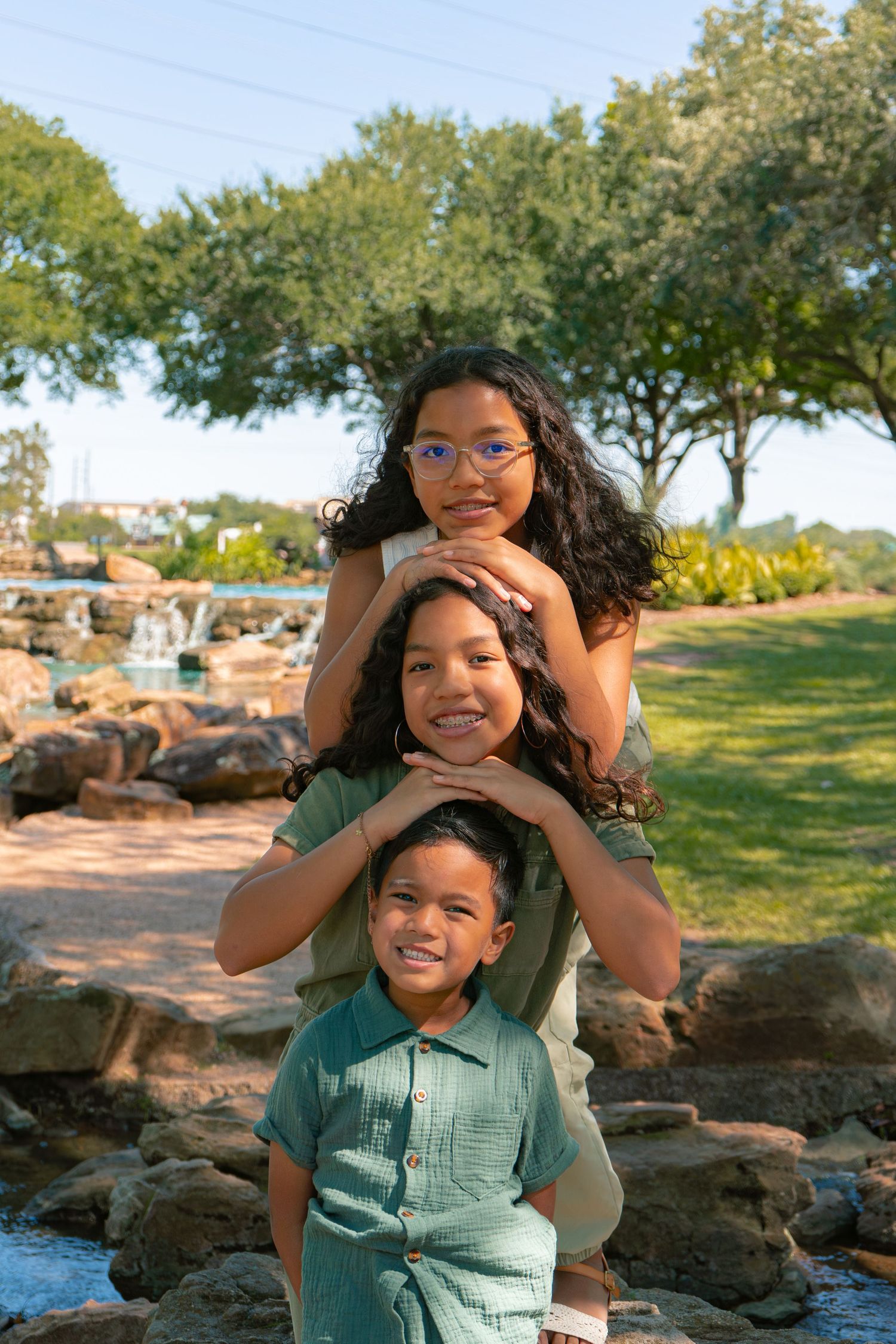Three children pose outdoors in a park, stacking hands on shoulders, smiling near a decorative fountain.