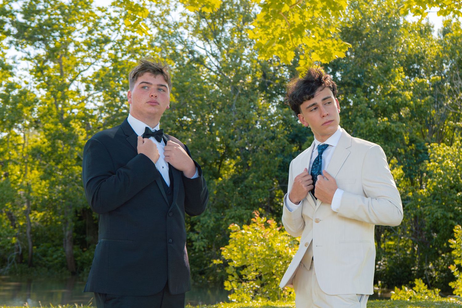 Two young men in formal suits adjust their ties while posing outdoors surrounded by lush green trees.