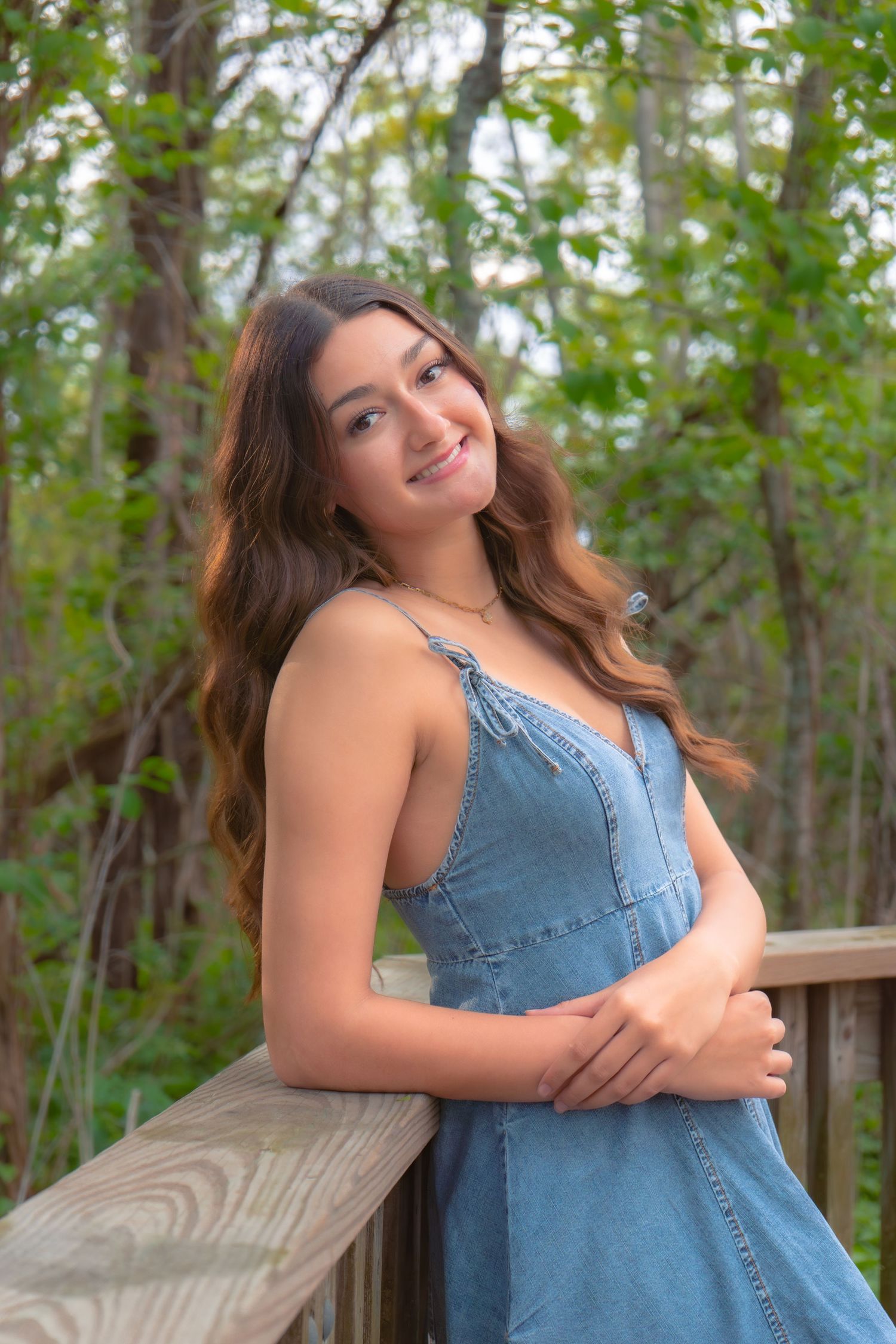 Young woman in denim sundress smiling on wooden deck surrounded by lush green trees.