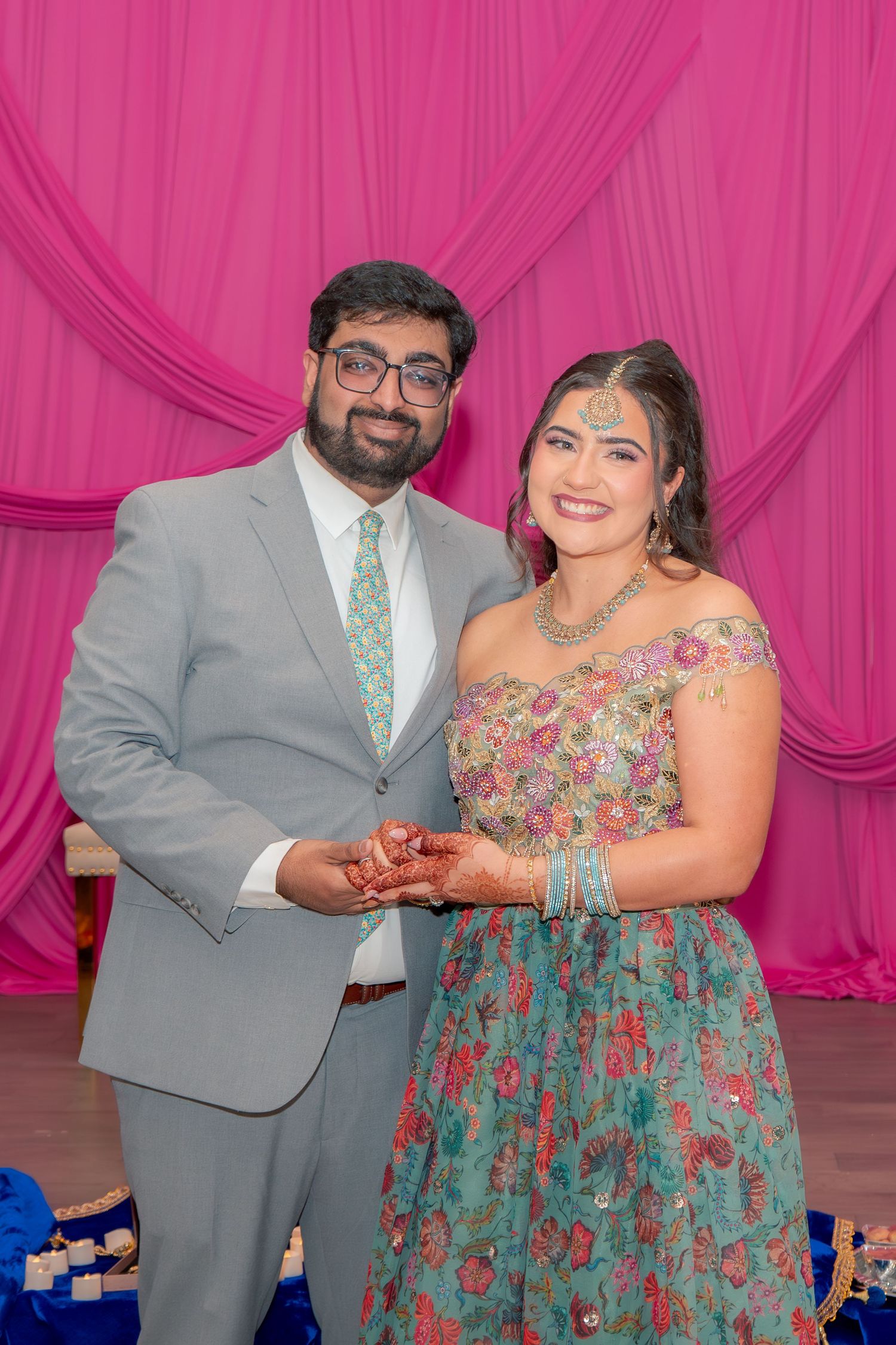 South Asian couple in traditional attire holding hands at a pink draped event backdrop.