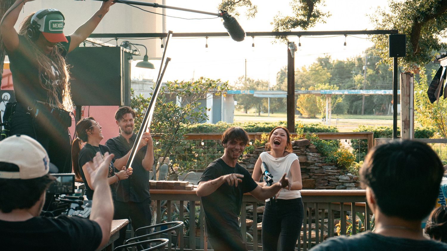 A film crew captures a lively outdoor scene on a sunny deck with actors laughing.