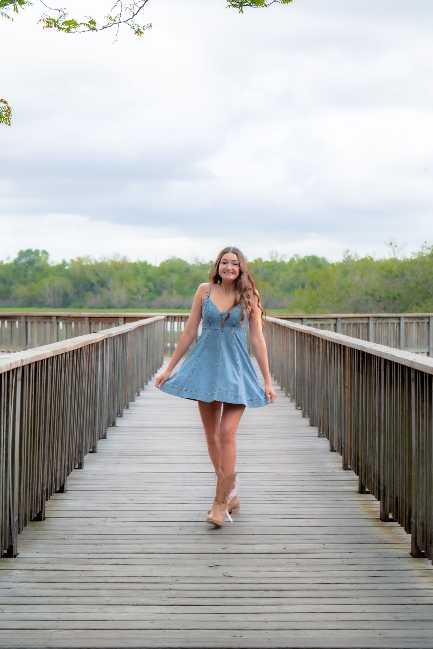 Smiling young woman in light blue dress walking along wooden boardwalk with green trees behind.