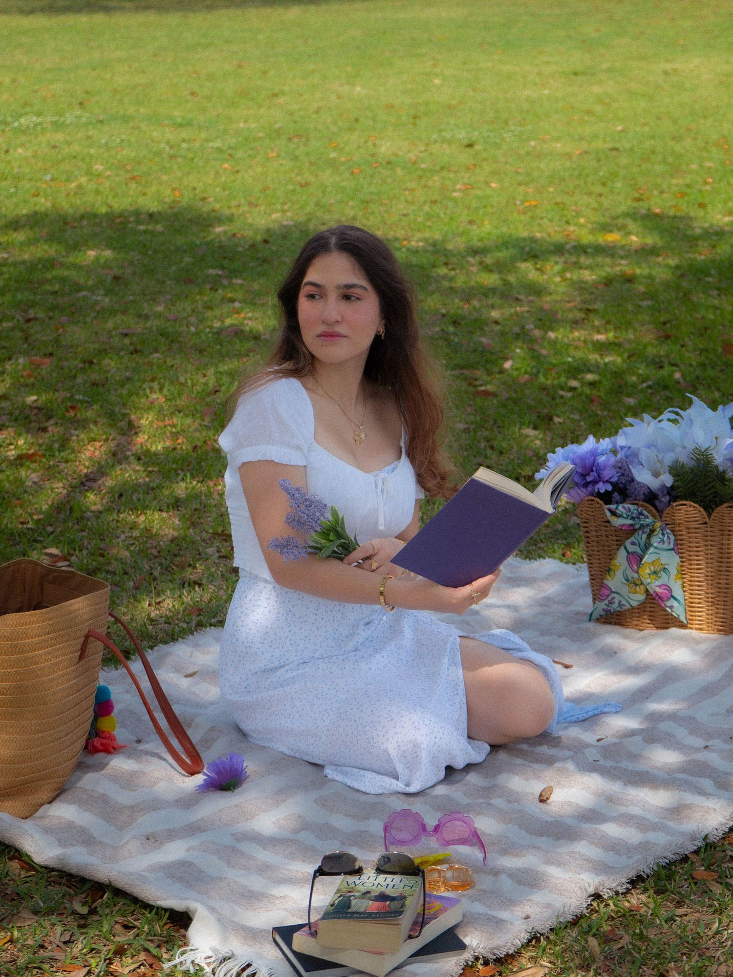 Young woman in white dress sitting on picnic blanket in park holding purple book and lavender flowers.