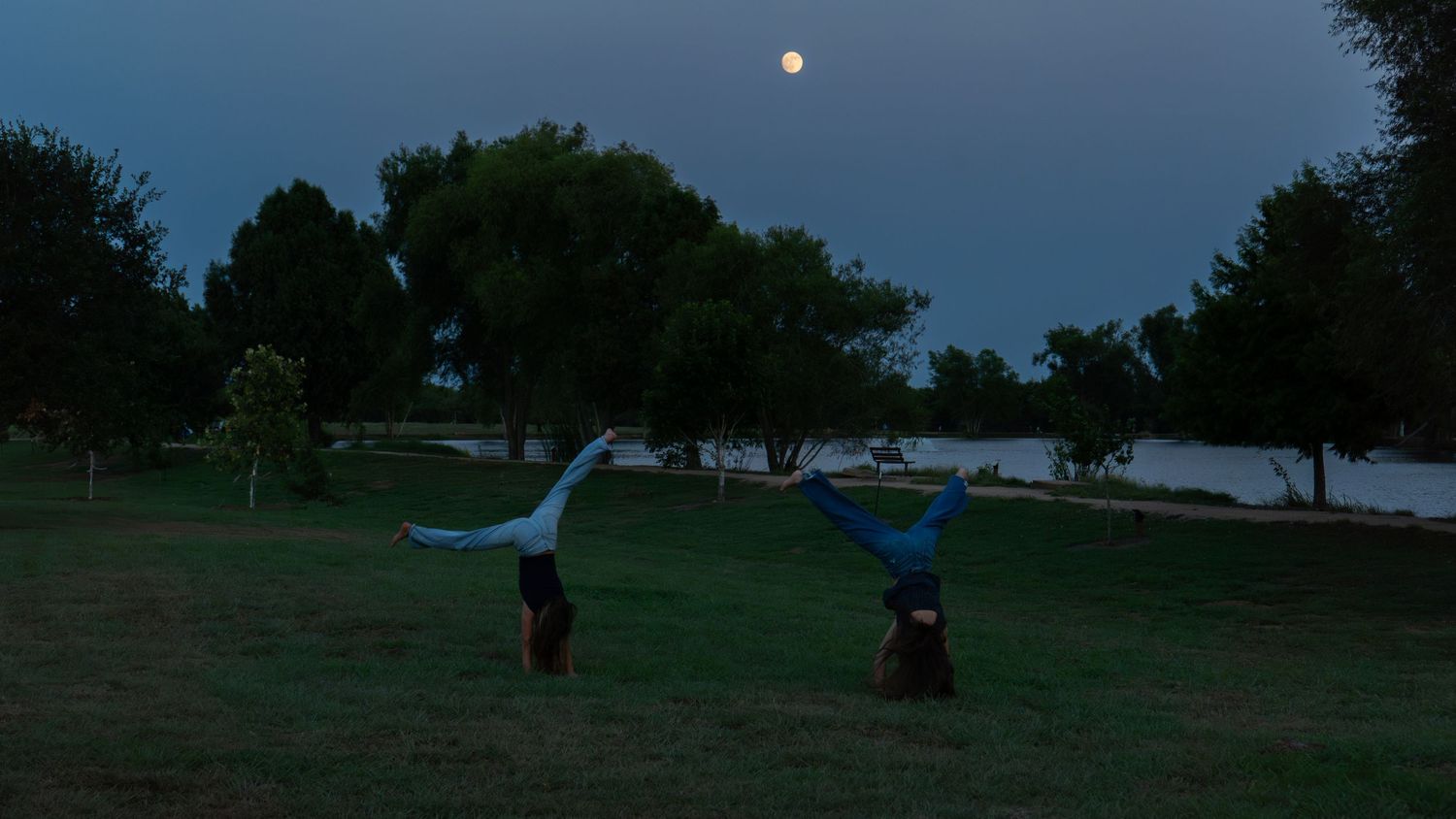 Two people doing handstands in a park at dusk with a full moon visible in the twilight sky.