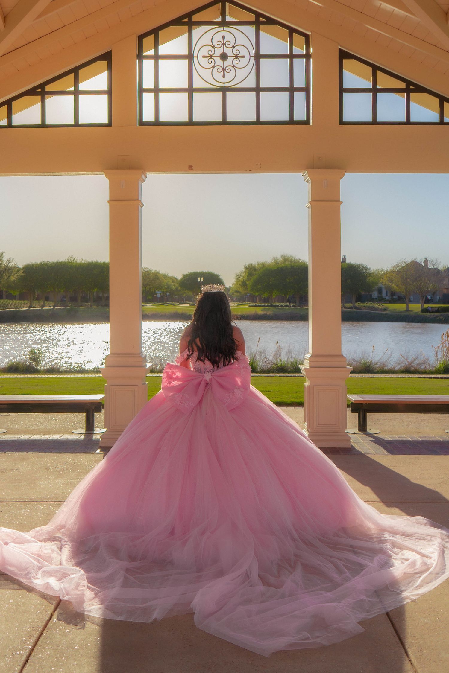 Young woman in a voluminous pink ball gown with bow stands in an elegant pavilion overlooking a serene lake.