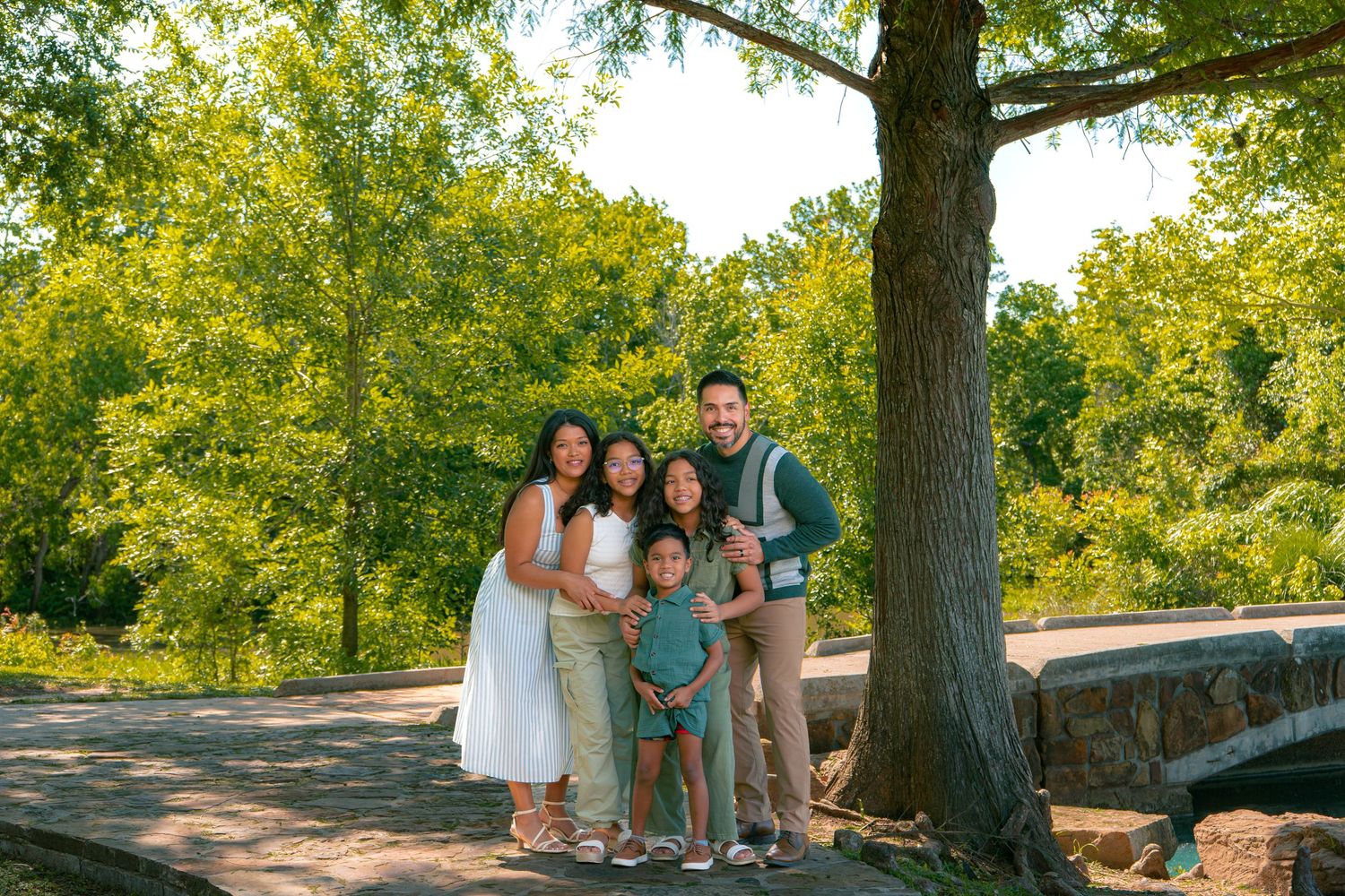 Happy family of five posing together outdoors near a large tree in a lush green park on a sunny day.