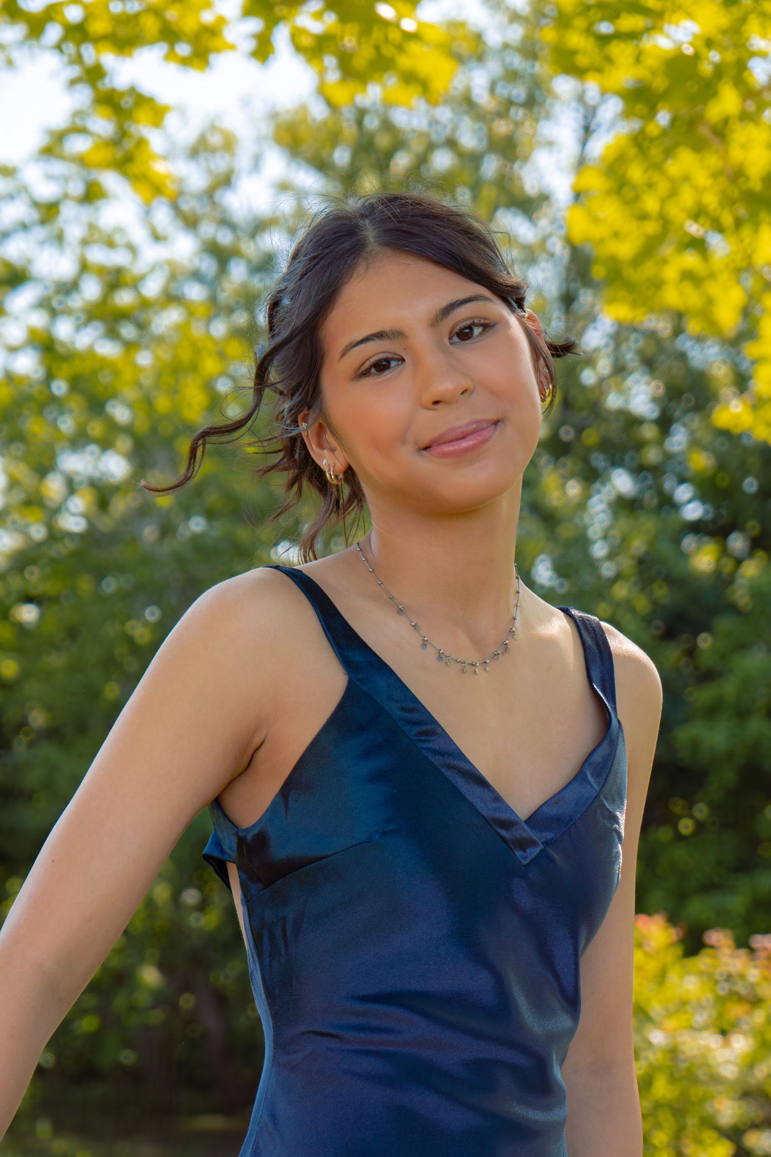 Young woman in a navy blue satin prom dress smiles softly outdoors with golden sunlit trees behind her.