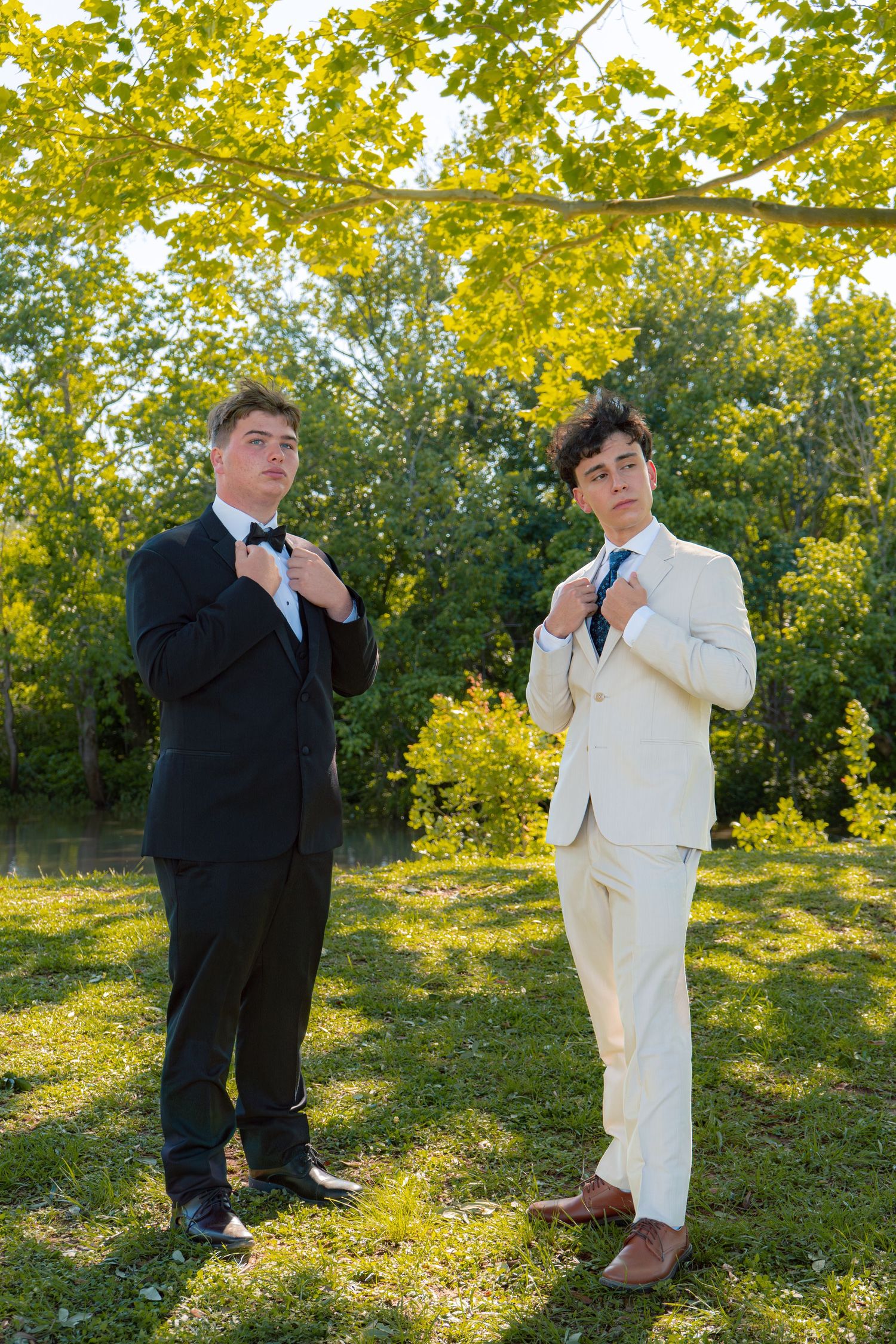 Two teenage boys in black tuxedo and cream suit adjust their ties outdoors under leafy tree branches.