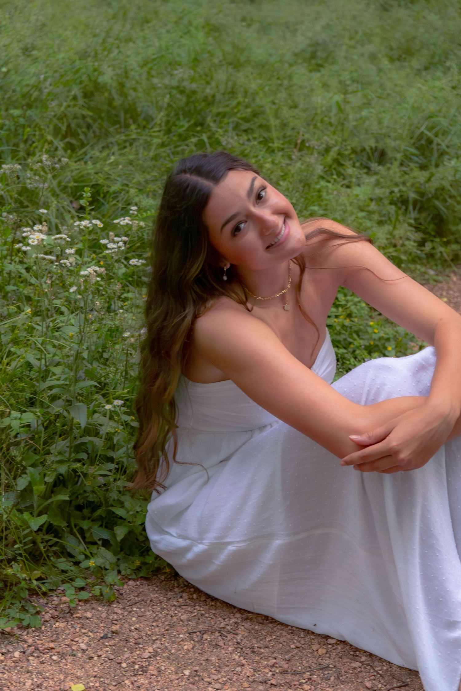 Young woman in white dress sitting on gravel path surrounded by lush green wildflowers, smiling softly.
