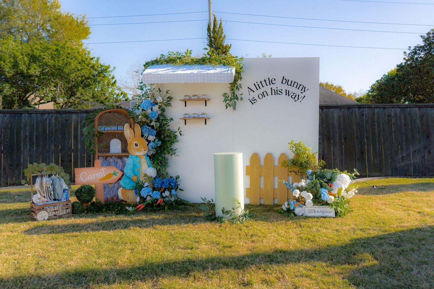 Peter Rabbit baby shower backdrop with balloon arch, white fence, and A Little Bunny Is On His Way sign.