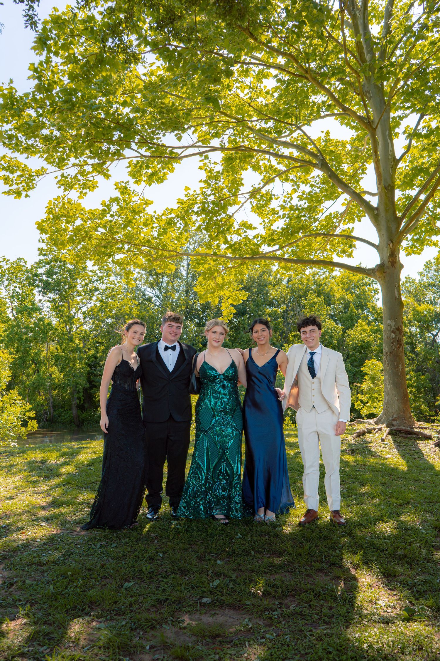 Five teenagers in formal prom attire pose outdoors under a large tree on a sunny day.