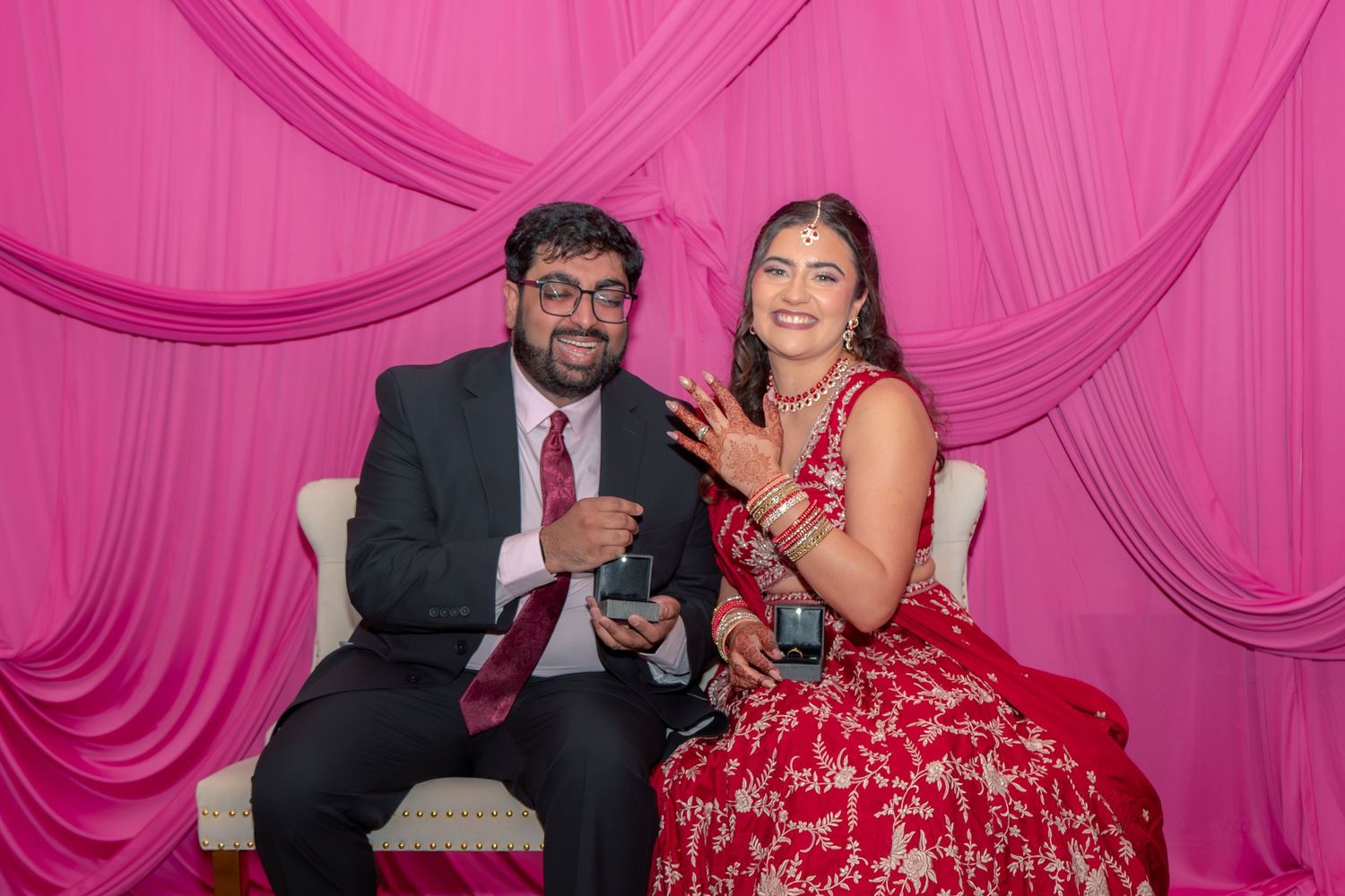 Couple in traditional Indian wedding attire smiling and showing rings against pink draped backdrop.