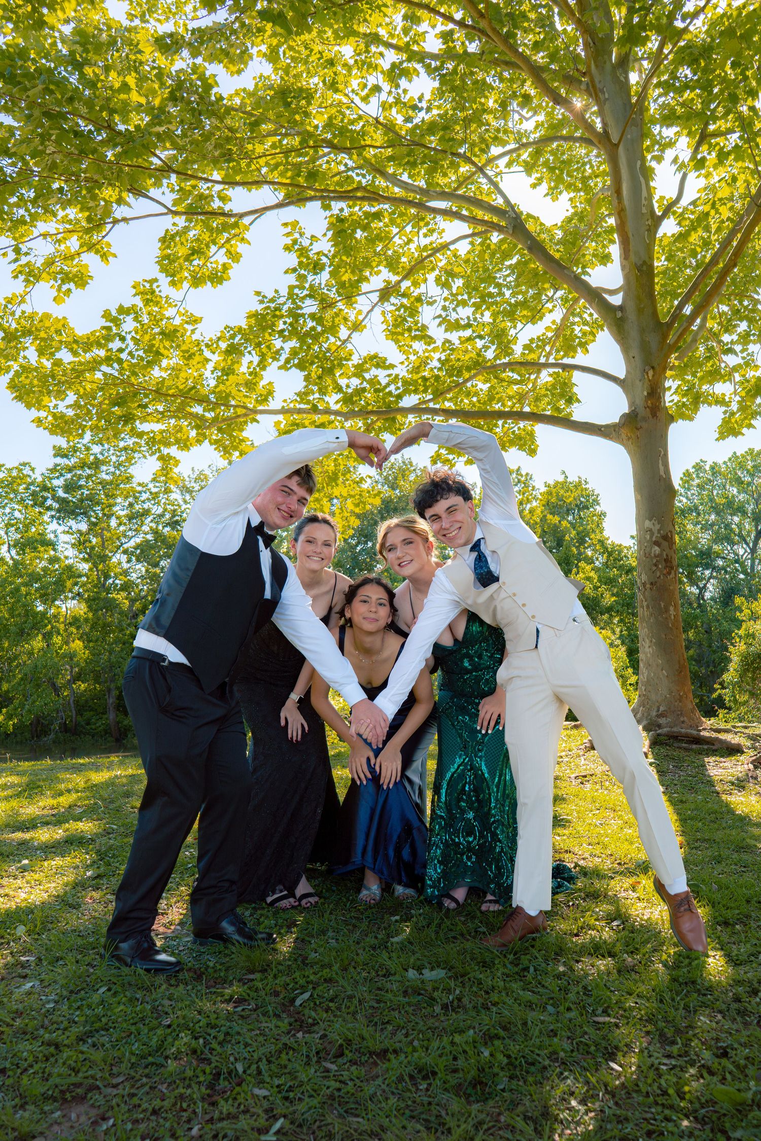 Group of five young people in formal prom attire form a heart shape with their arms under a large tree.