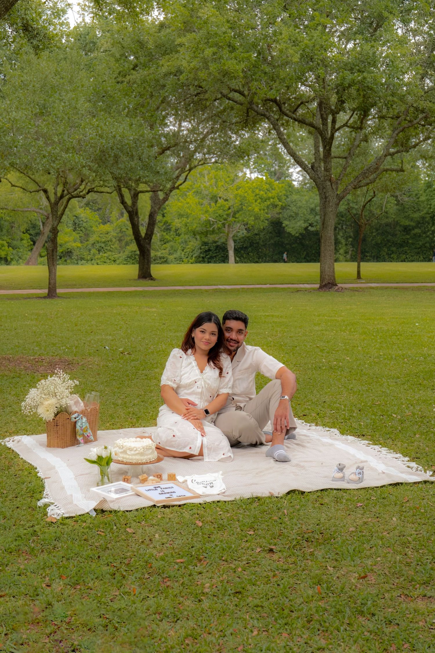 Couple sitting together on a white blanket in a lush green park, celebrating with a cake and picnic setup.