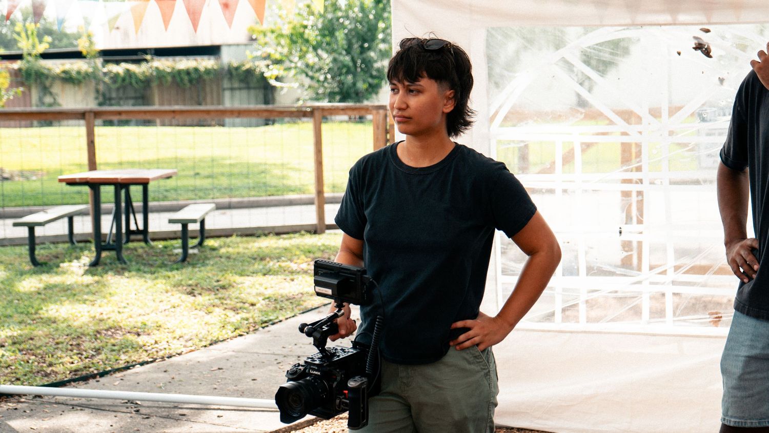 A filmmaker holds a professional camera on a sunny outdoor set with a gazebo and picnic table in the background.