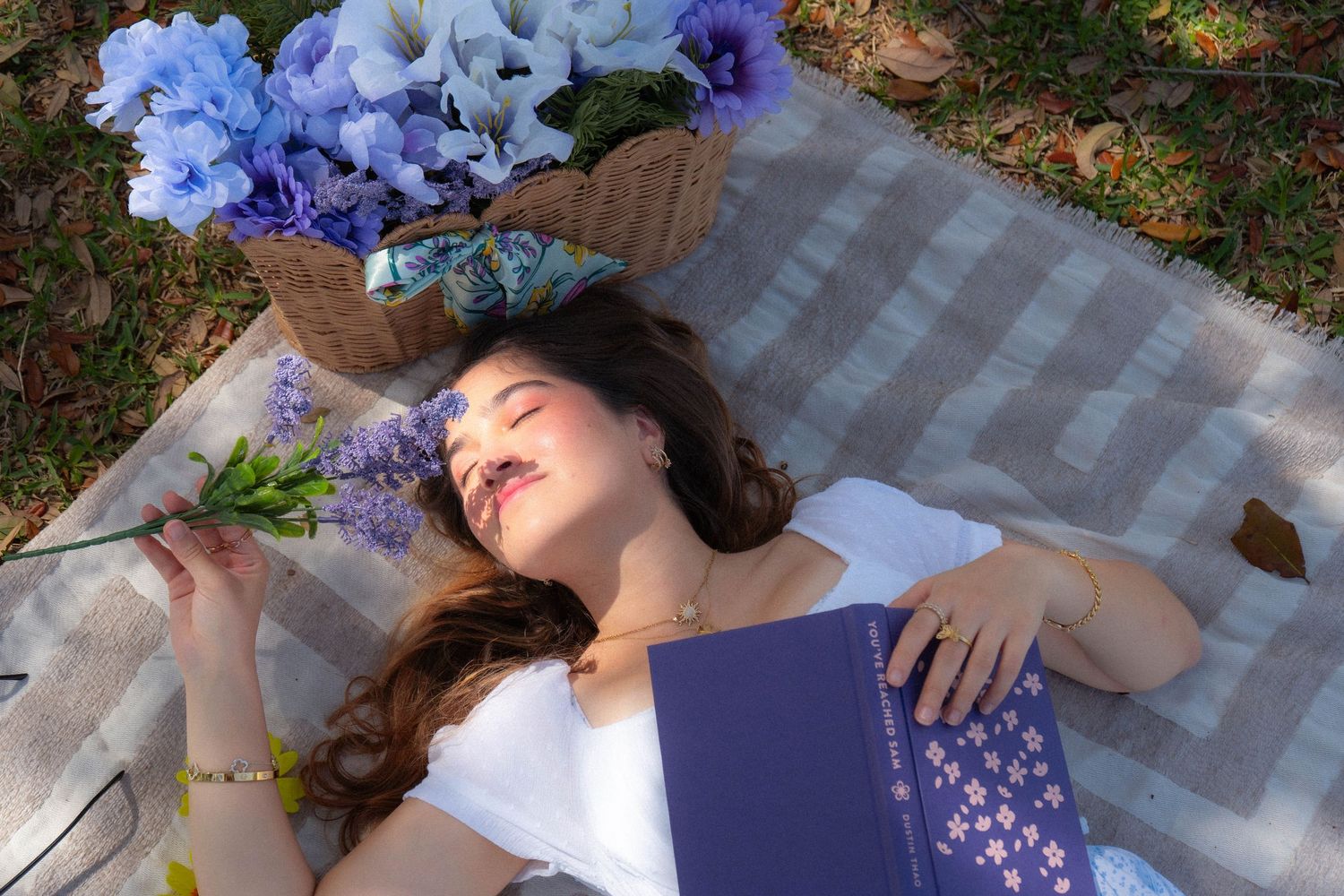Peaceful woman resting on picnic blanket with blue book, holding purple flowers near wicker basket of blue florals.