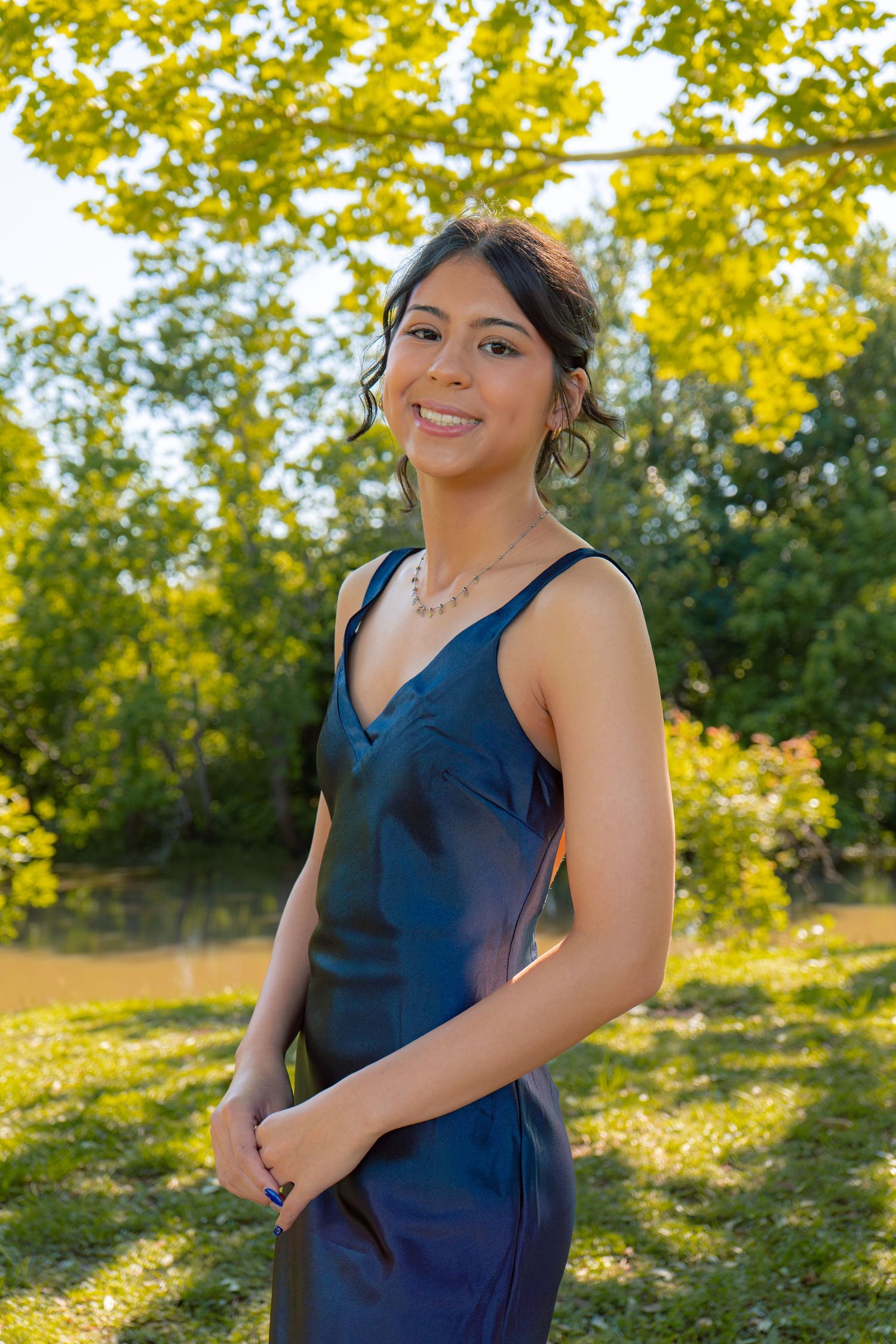 Smiling young woman in a navy blue formal gown wearing a pearl necklace poses outdoors among green trees.
