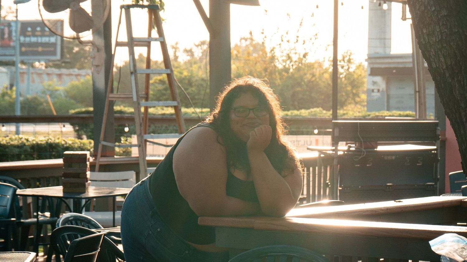 Woman wearing sunglasses relaxes at an outdoor patio table in warm golden sunlight.