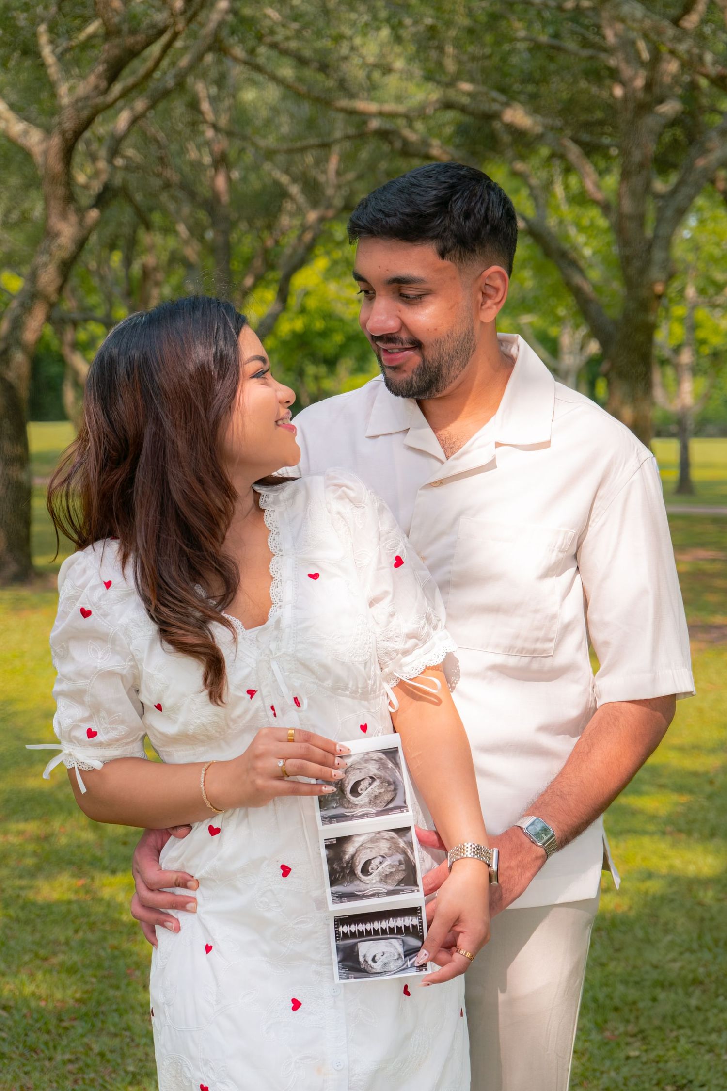 Smiling couple in white outfits holding ultrasound photos in a park, announcing their pregnancy.