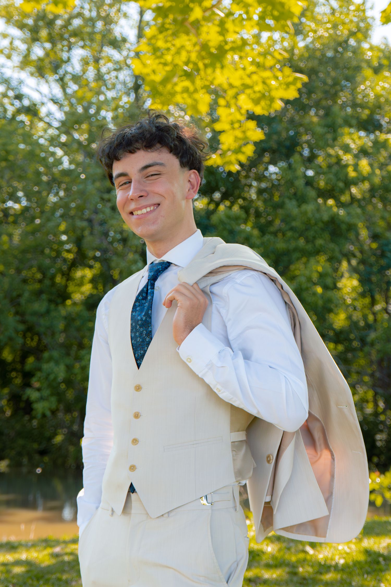 Stylish young man in a cream three-piece suit smiles while holding his jacket over his shoulder outdoors in sunlight.