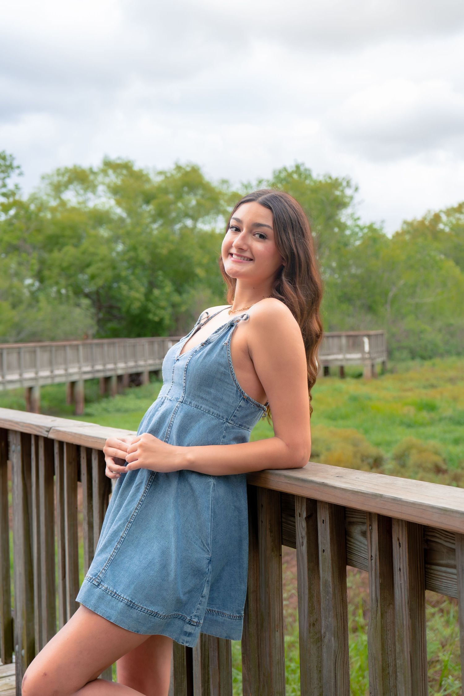 Cheerful young woman in denim sundress leaning against wooden railing on outdoor boardwalk nature trail.