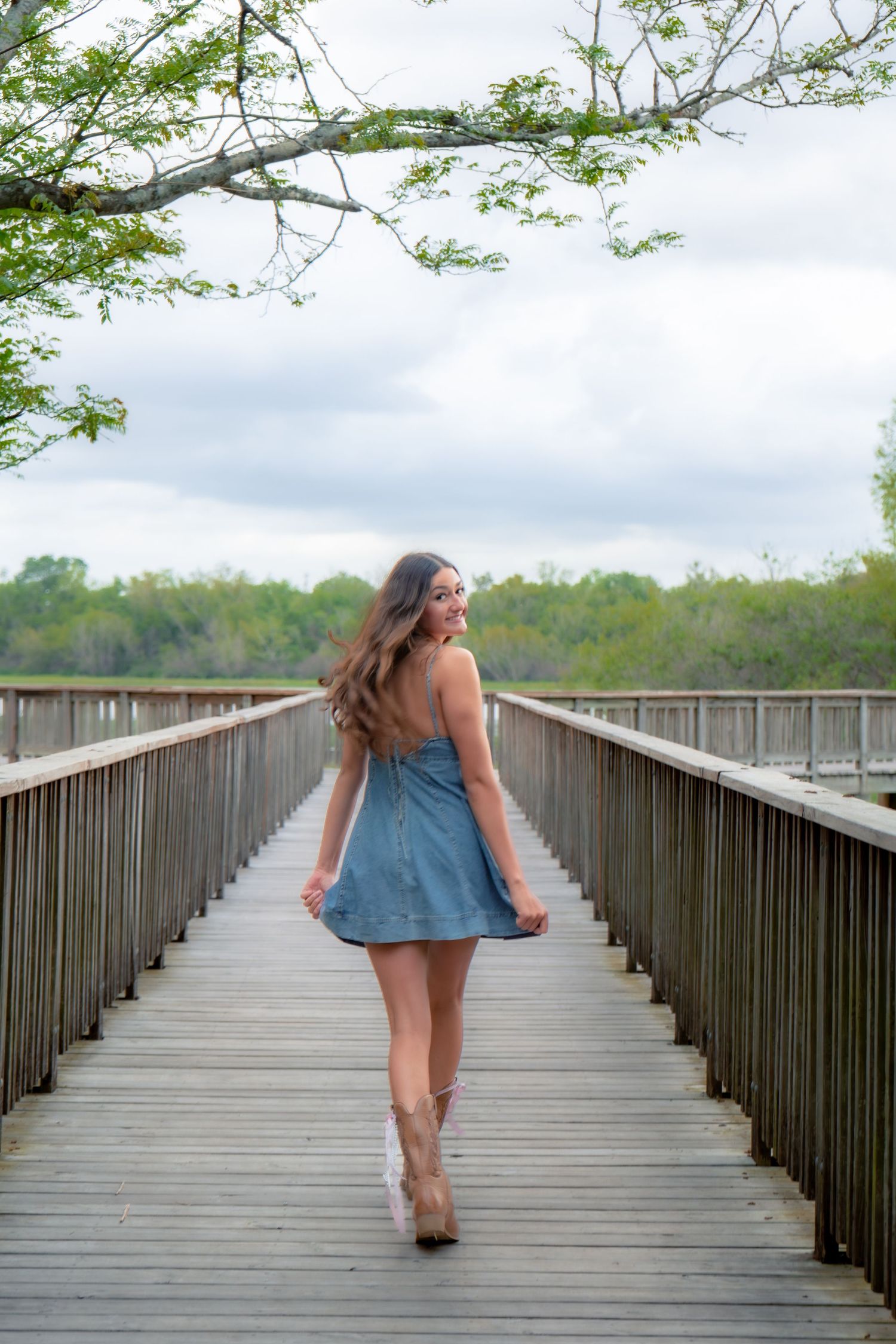 Young woman in denim dress walking on wooden boardwalk through nature preserve.