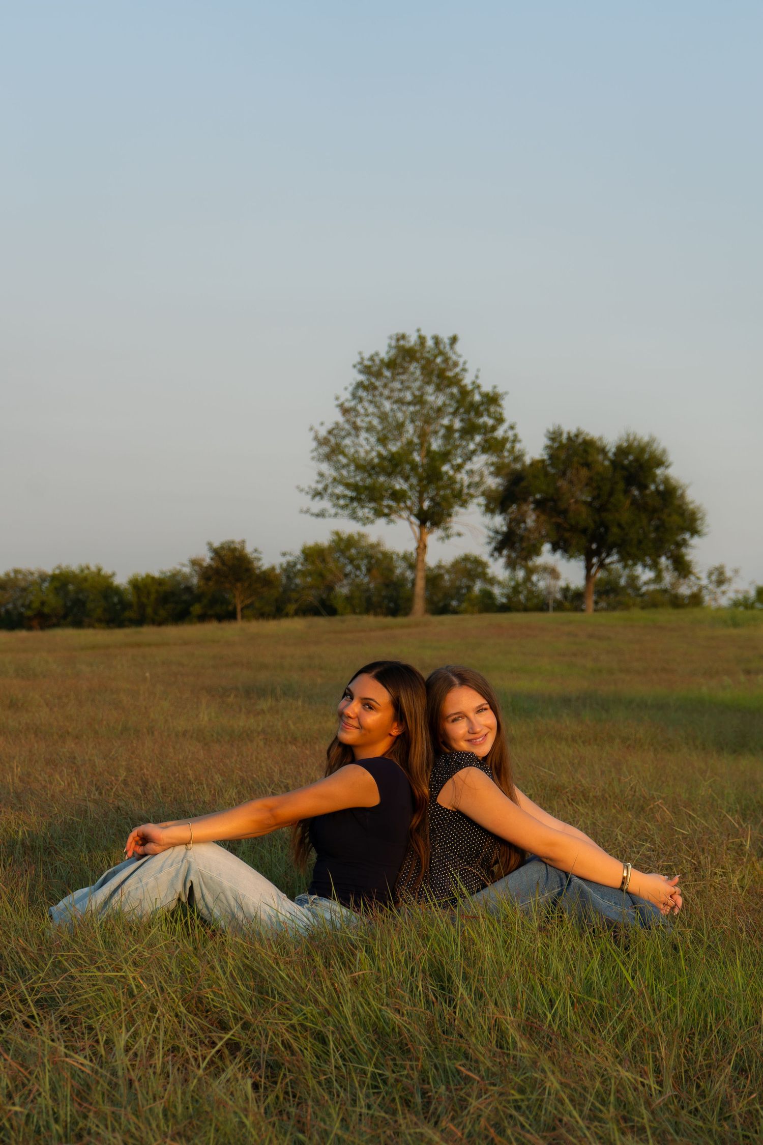 Two young women sitting back-to-back in a grassy field, smiling, with trees and blue sky in background.