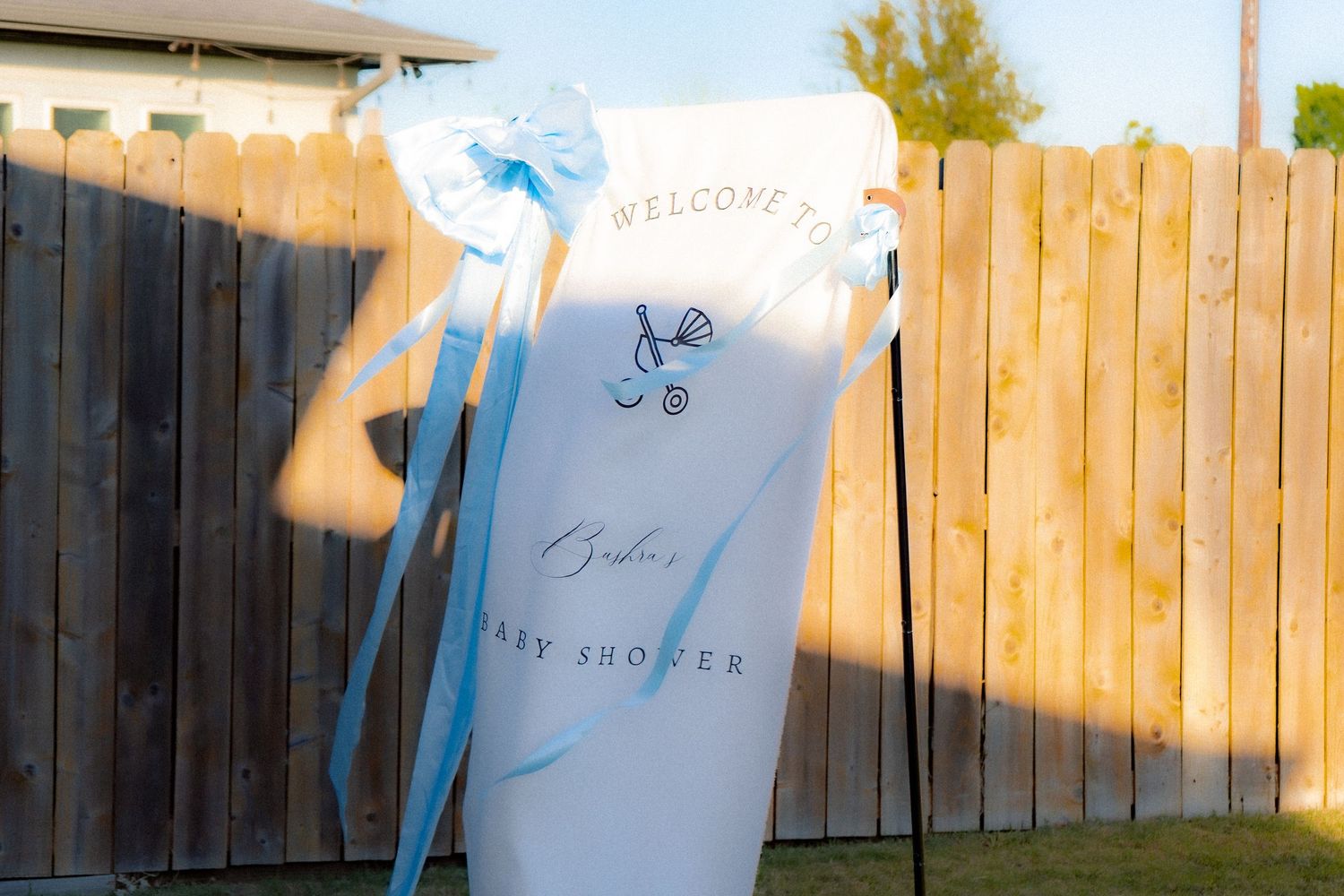 Outdoor baby shower welcome banner with blue bow decorations and baby stroller graphic against a wooden fence backdrop.