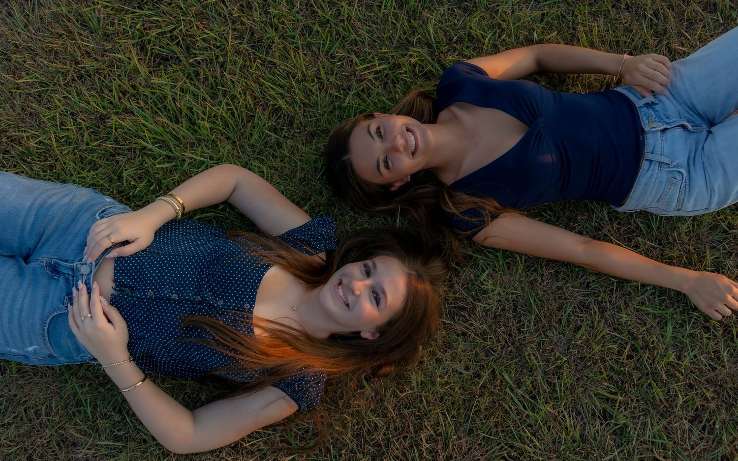 Two young women lying on green grass, smiling and relaxing outdoors in casual jeans and blue tops.