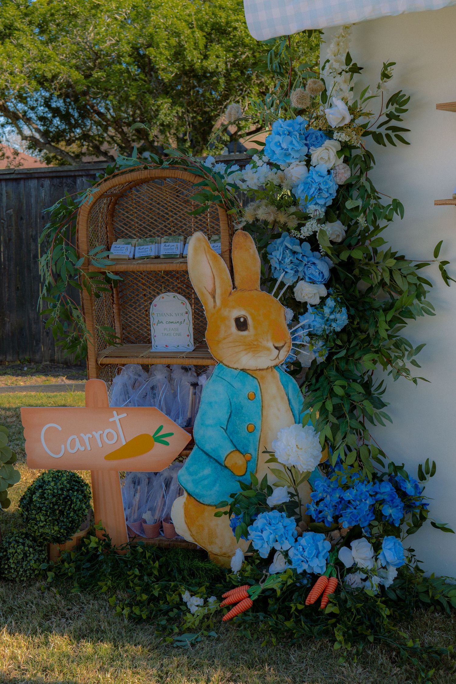 Peter Rabbit character cutout with carrot sign, blue hydrangeas, white flowers, and wicker shelf decoration.