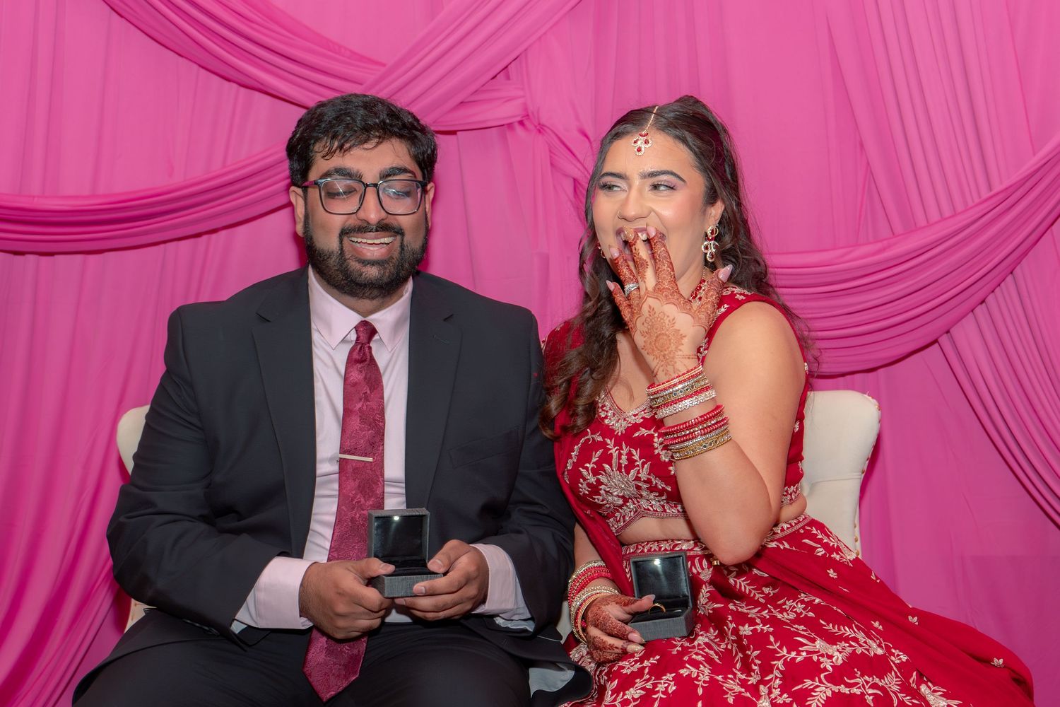 Indian couple at wedding ceremony, woman in red bridal lehenga showing mehndi hands, man in dark suit beside her.