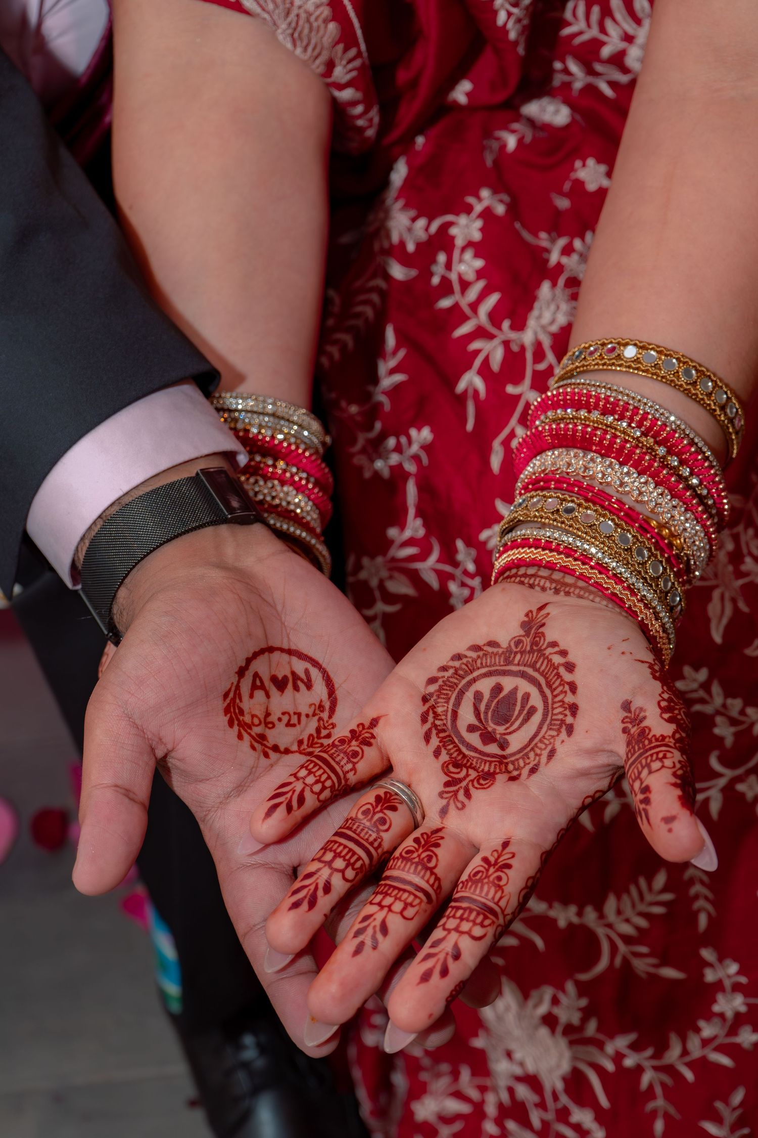 Bride displaying intricate red mehndi henna designs on palms with gold bangles and initials, beside groom in suit.