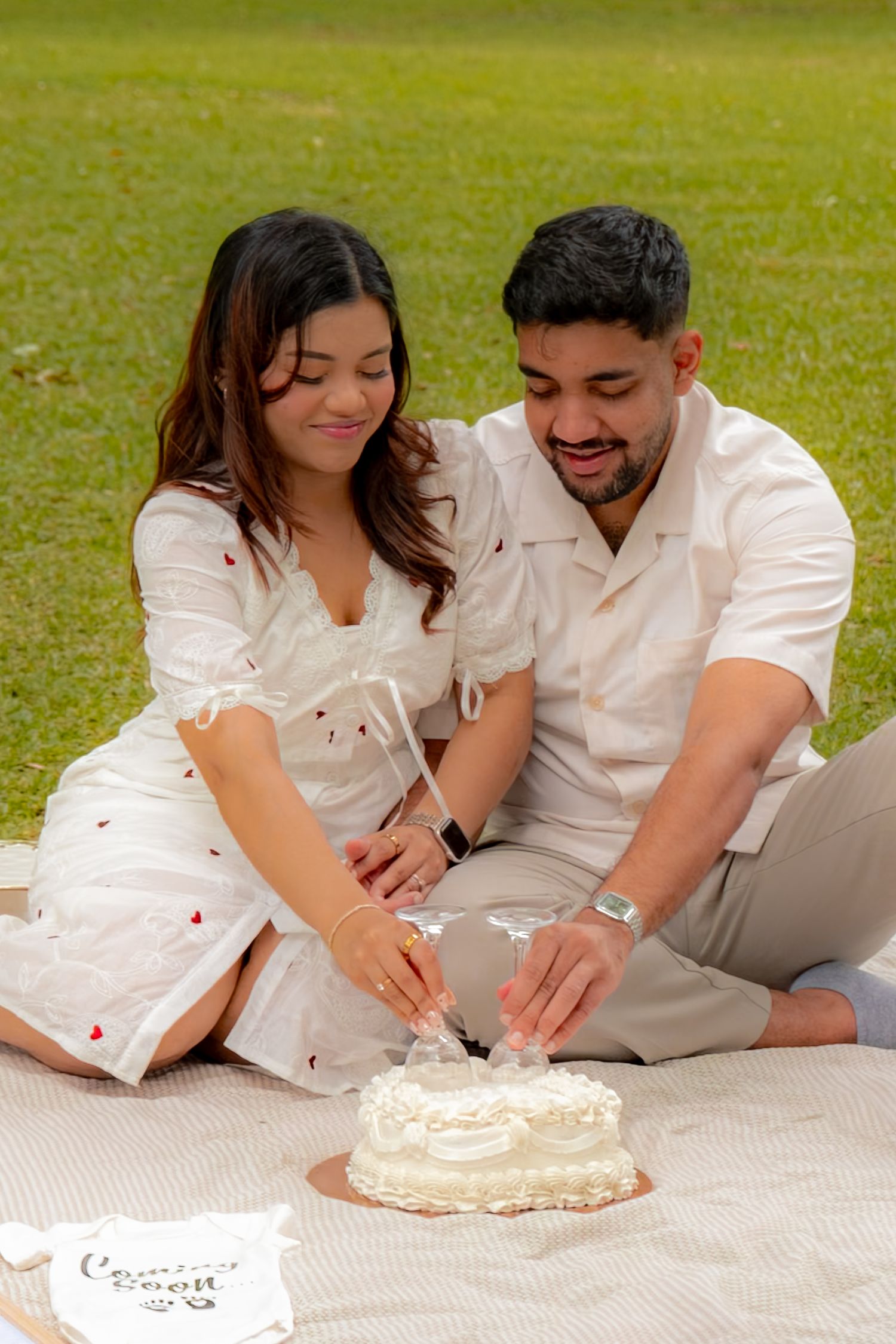 Smiling couple in white outfits cutting a decorated cake together on a picnic blanket outdoors.