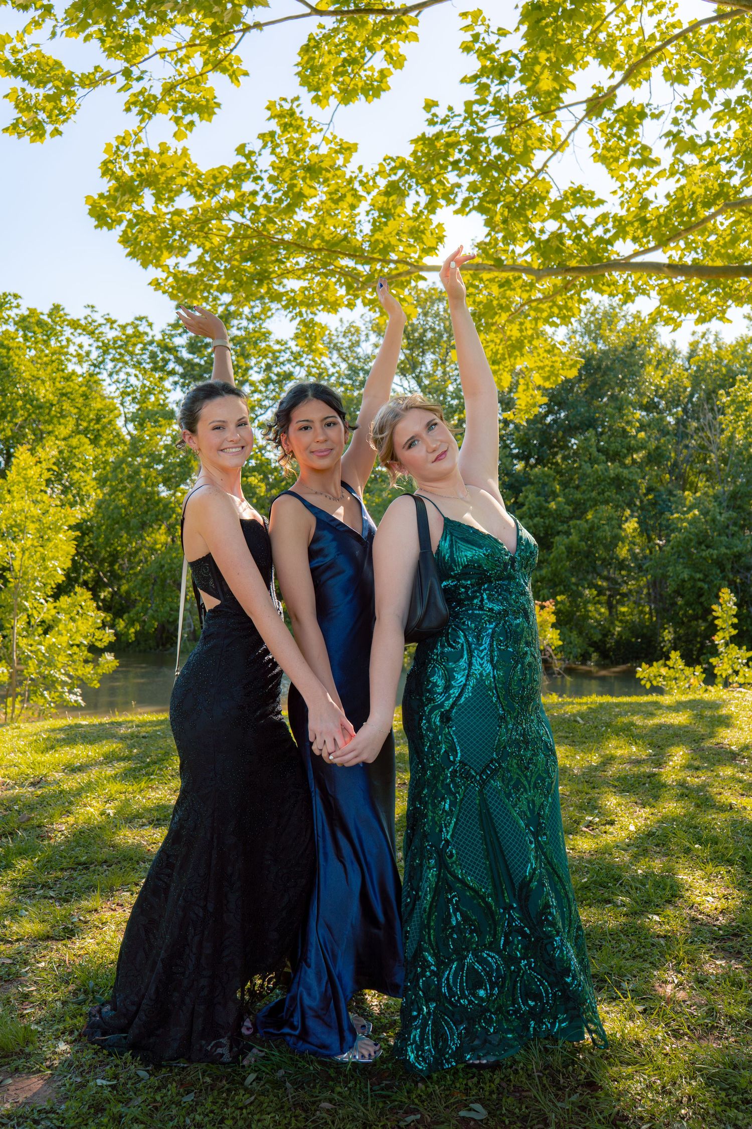 Three young women in black, navy, and green formal prom gowns posing together under tree branches outdoors.