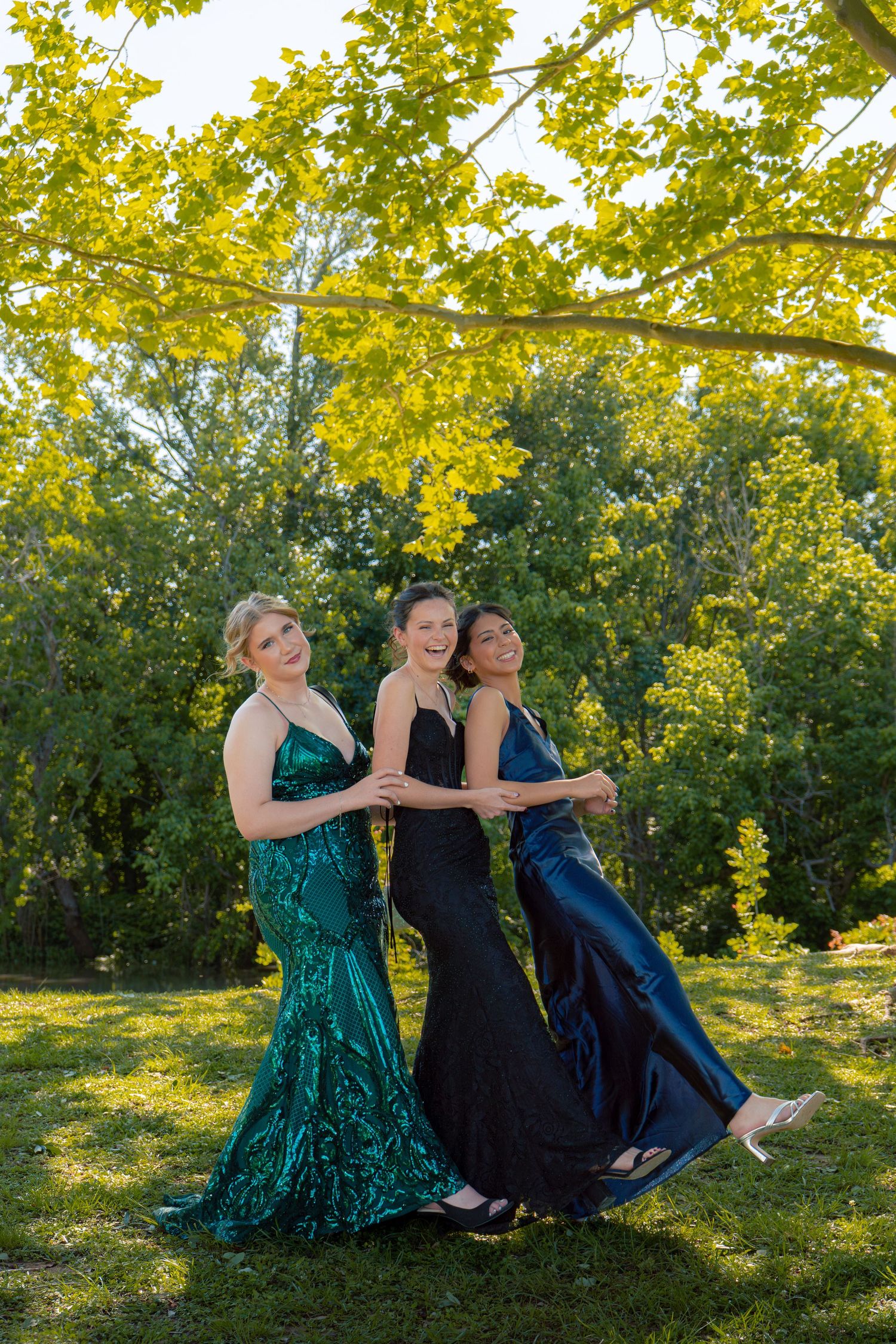 Three young women in elegant prom gowns pose playfully together on a sunny green lawn beneath yellow-leafed trees.