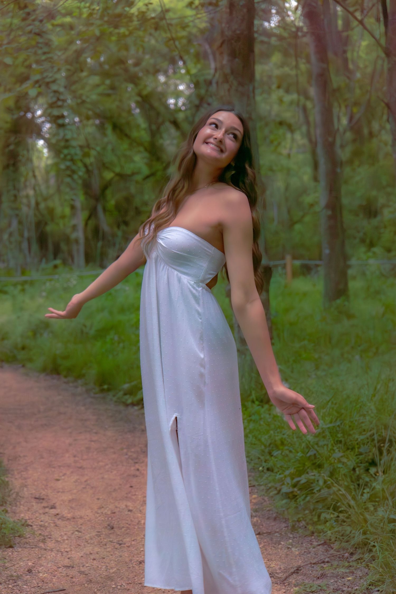 Young woman in white strapless maxi dress posing on forest path with arms outstretched.