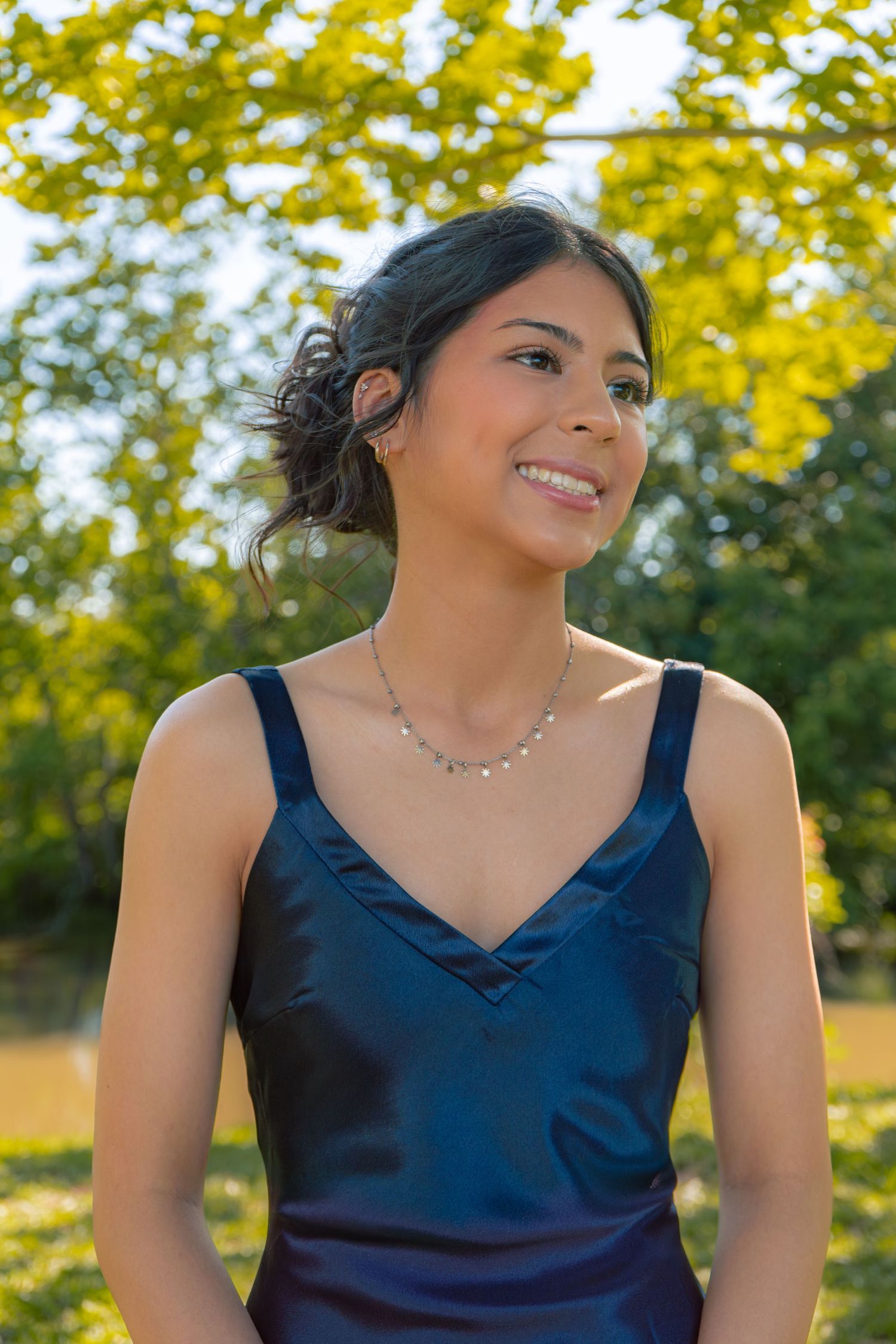 Young woman in navy blue satin dress smiling outdoors with lush green trees in background.