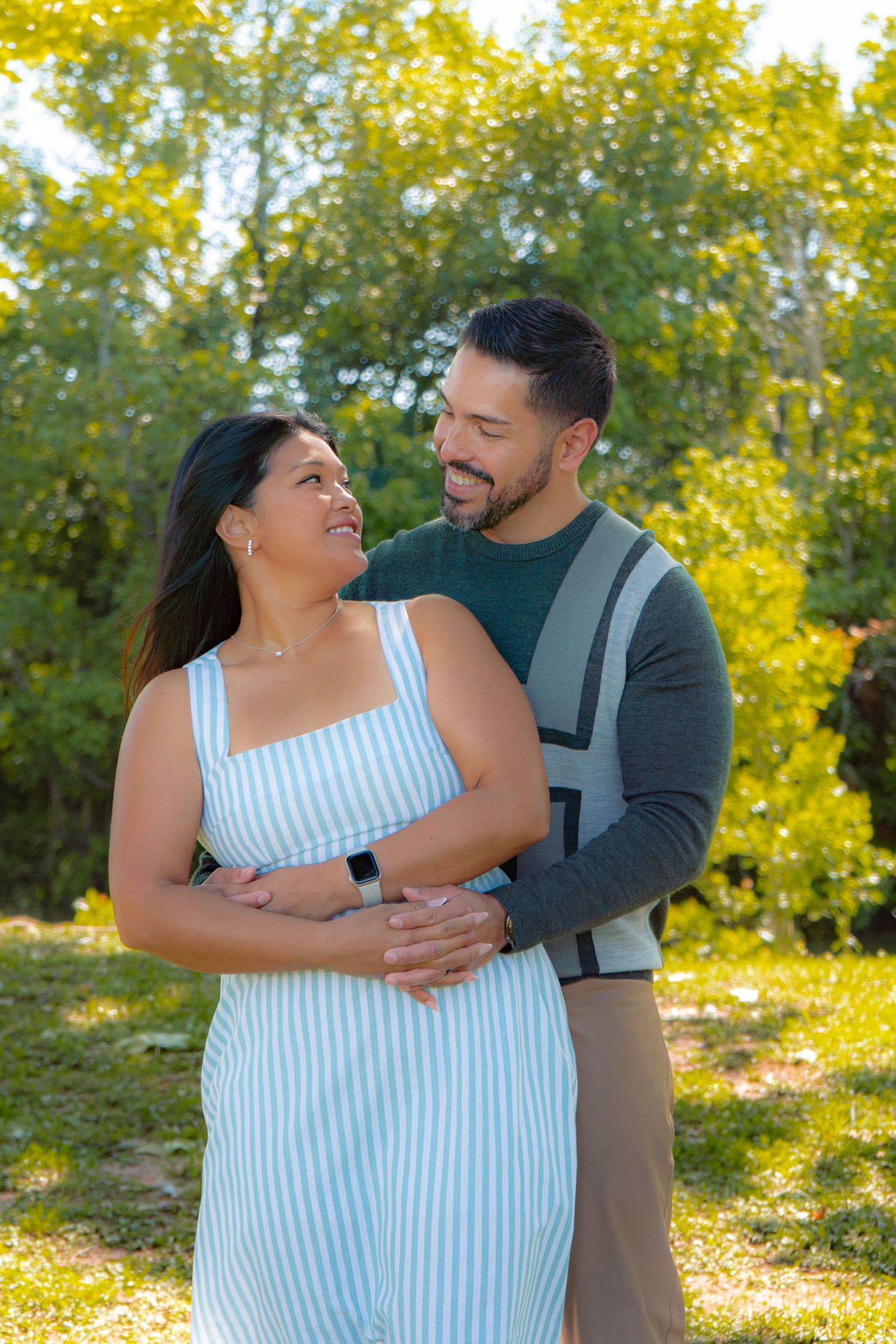A smiling couple embraces outdoors in a sunny park, woman in striped dress, man in green sweater with suspenders.