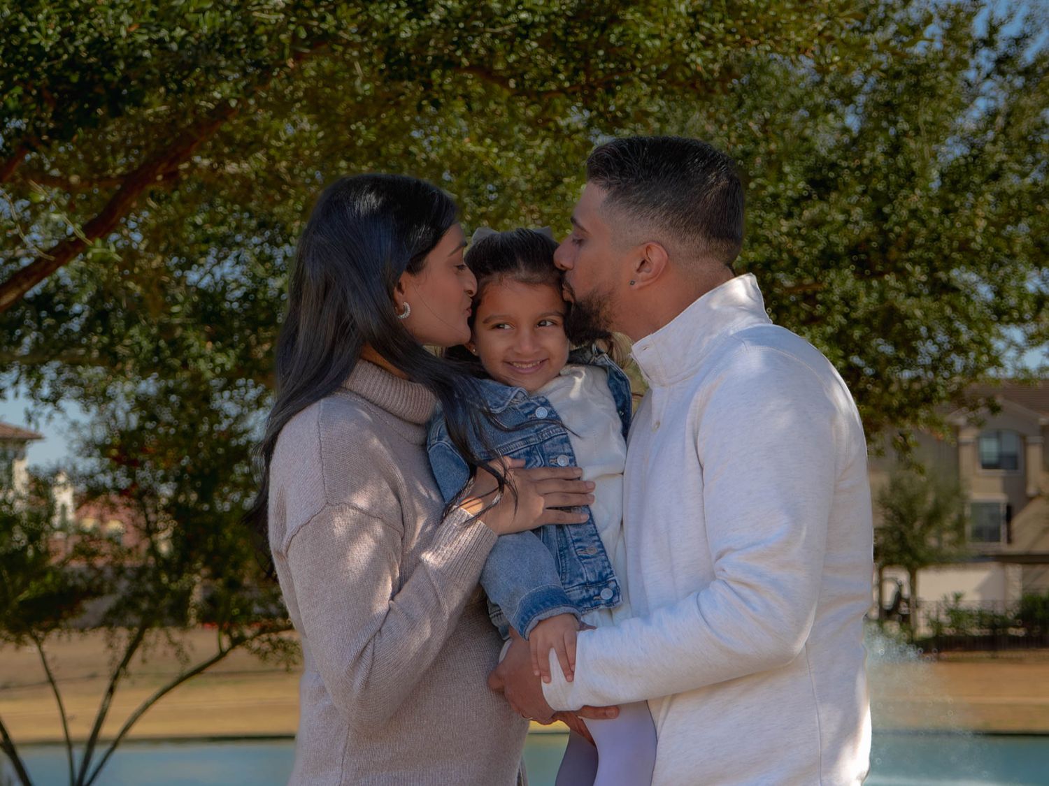 Loving parents in neutral tones kiss their smiling toddler daughter outdoors under green trees.