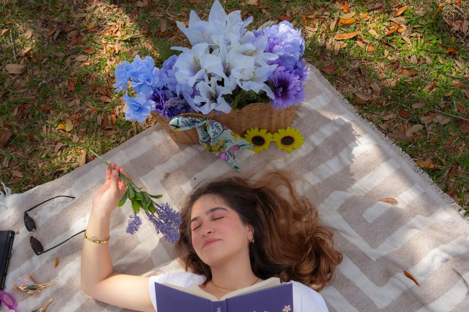 Young woman lying on blanket reading purple book, holding lavender flowers beside basket of blue and white blooms.