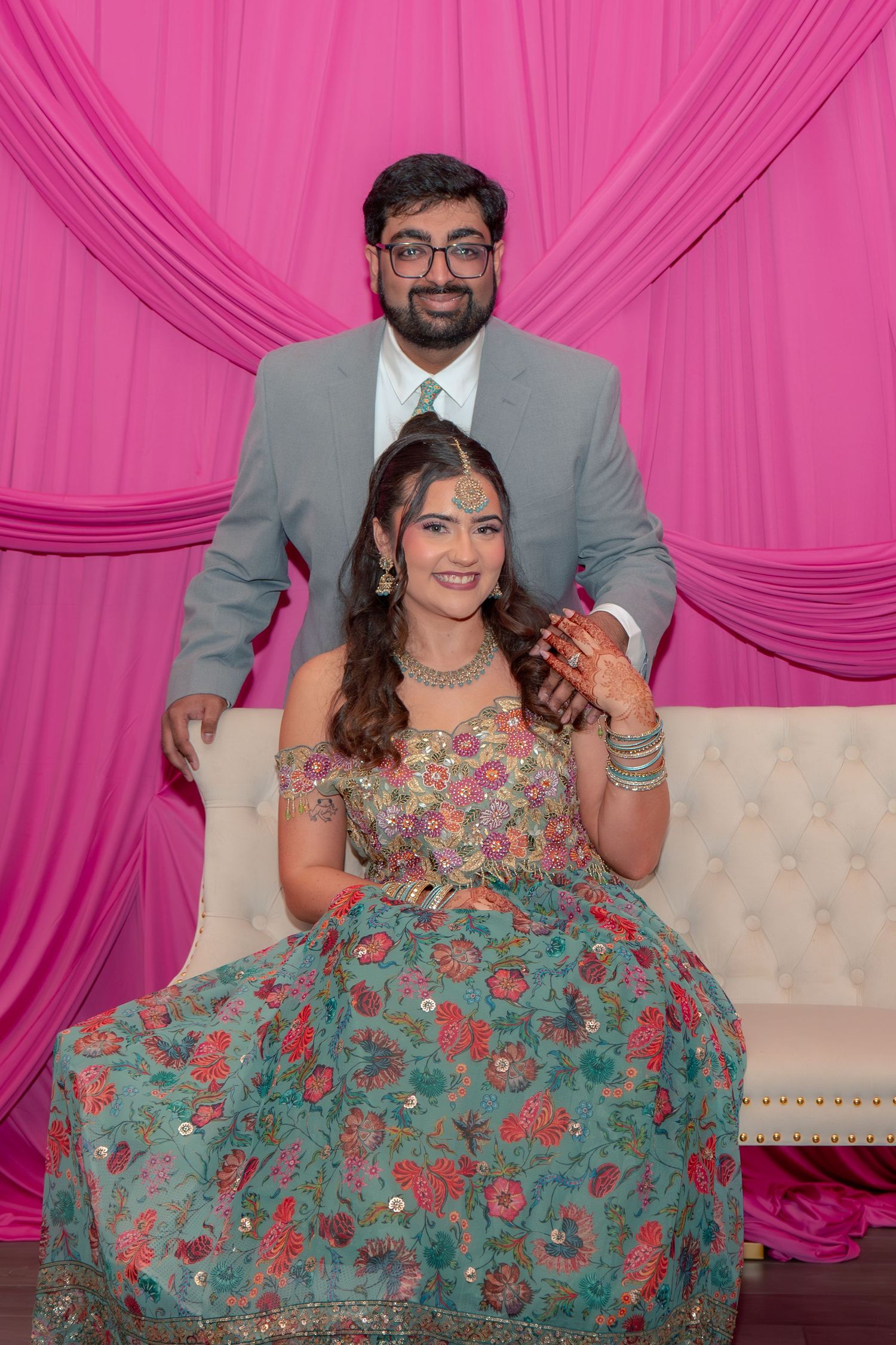 Indian couple posing at engagement ceremony, woman in floral lehenga seated on white couch, man in gray suit behind her.