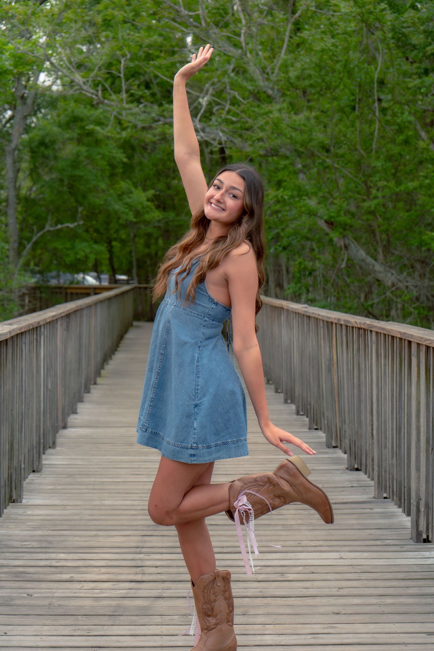 Young woman in denim dress and cowboy boots posing with arm raised on wooden boardwalk.