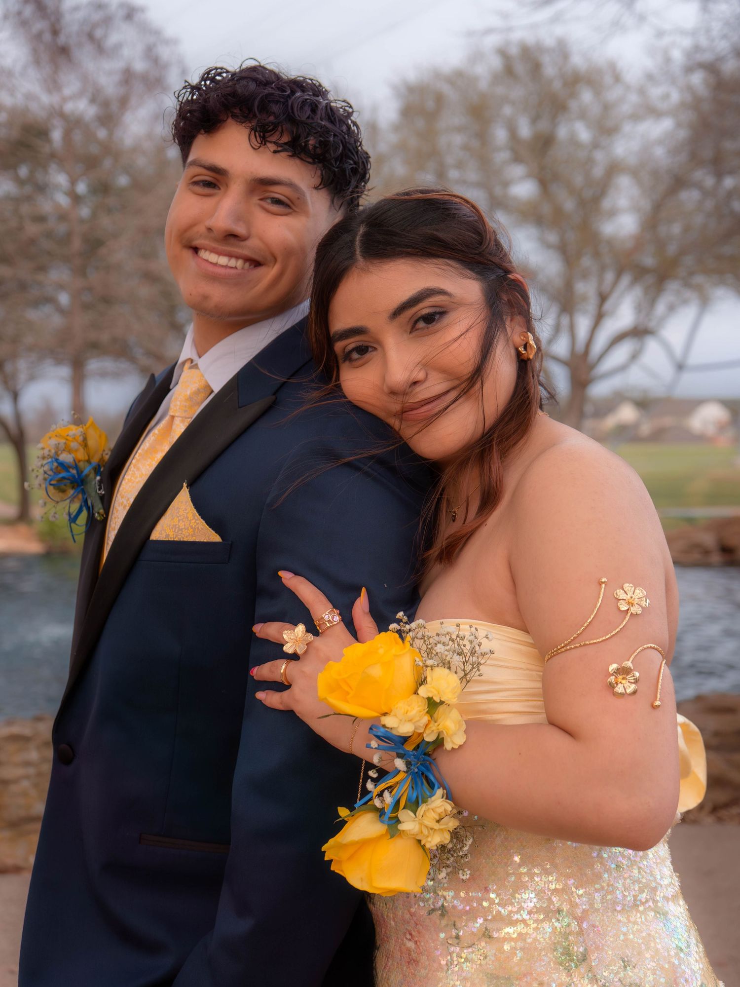 A smiling couple in prom attire, man in navy tuxedo with gold tie, woman in strapless gown with yellow floral corsages.