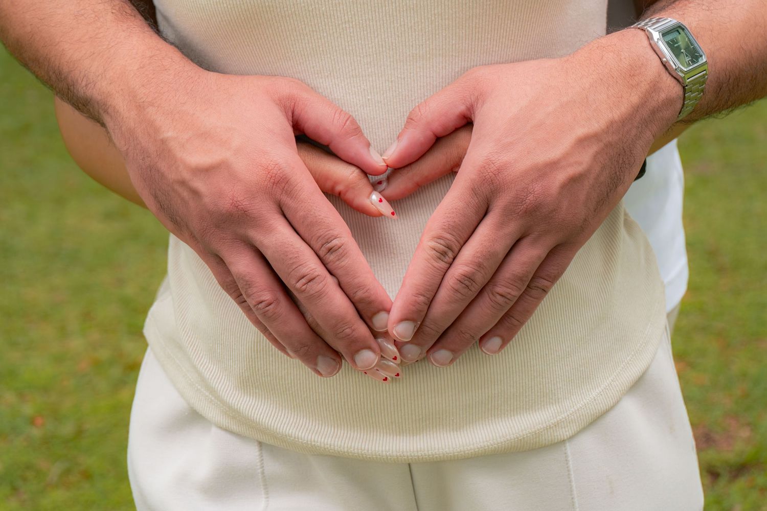 Couple forming a heart shape with hands on pregnant belly, wearing wedding rings outdoors.