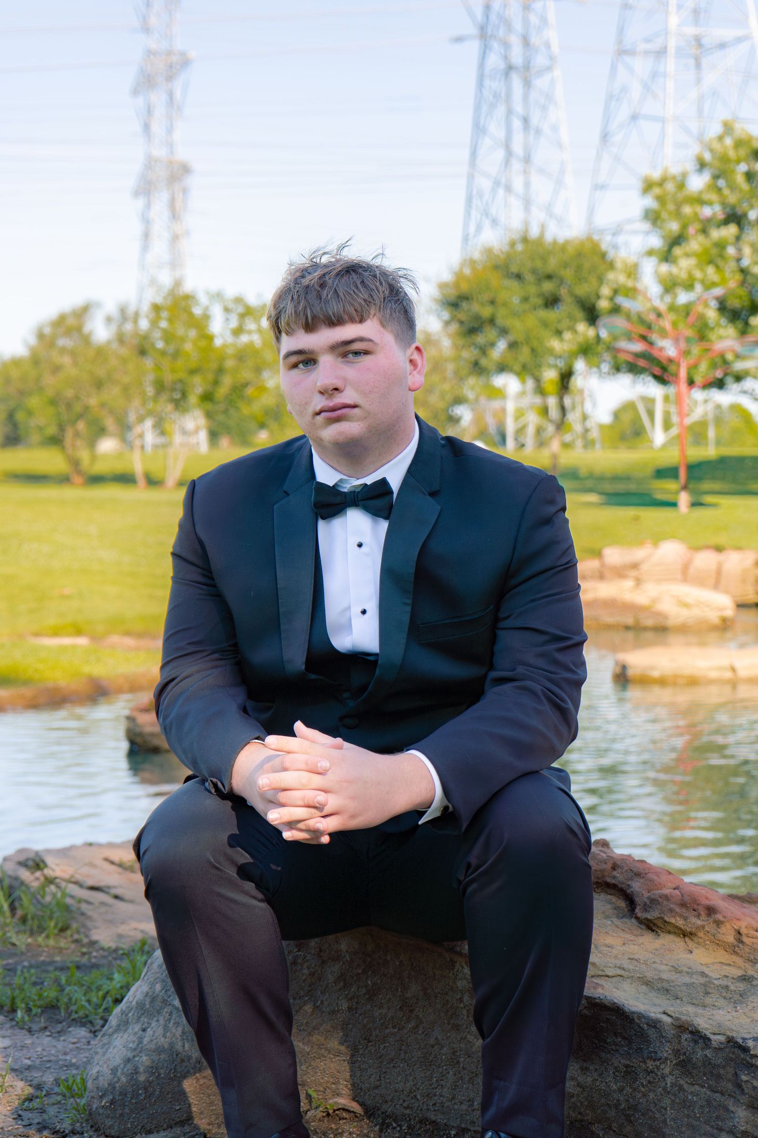 Young man in classic black tuxedo with bow tie sitting on rocks near pond at golden hour.