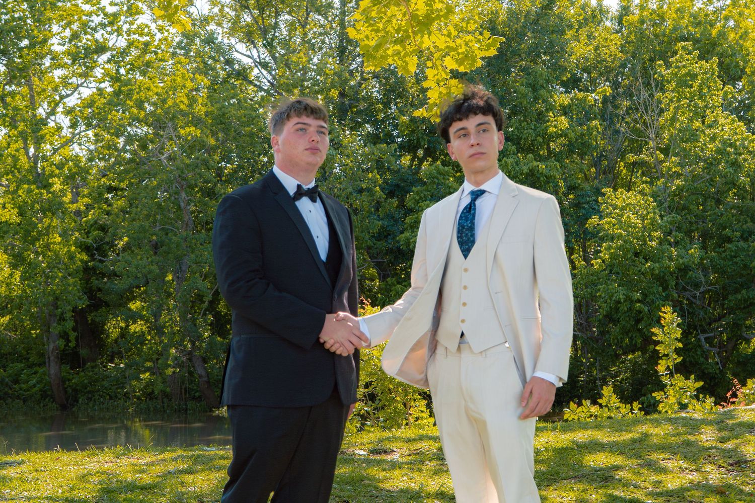 Two young men in formal suits shaking hands outdoors at prom, one in black tuxedo, one in white suit.