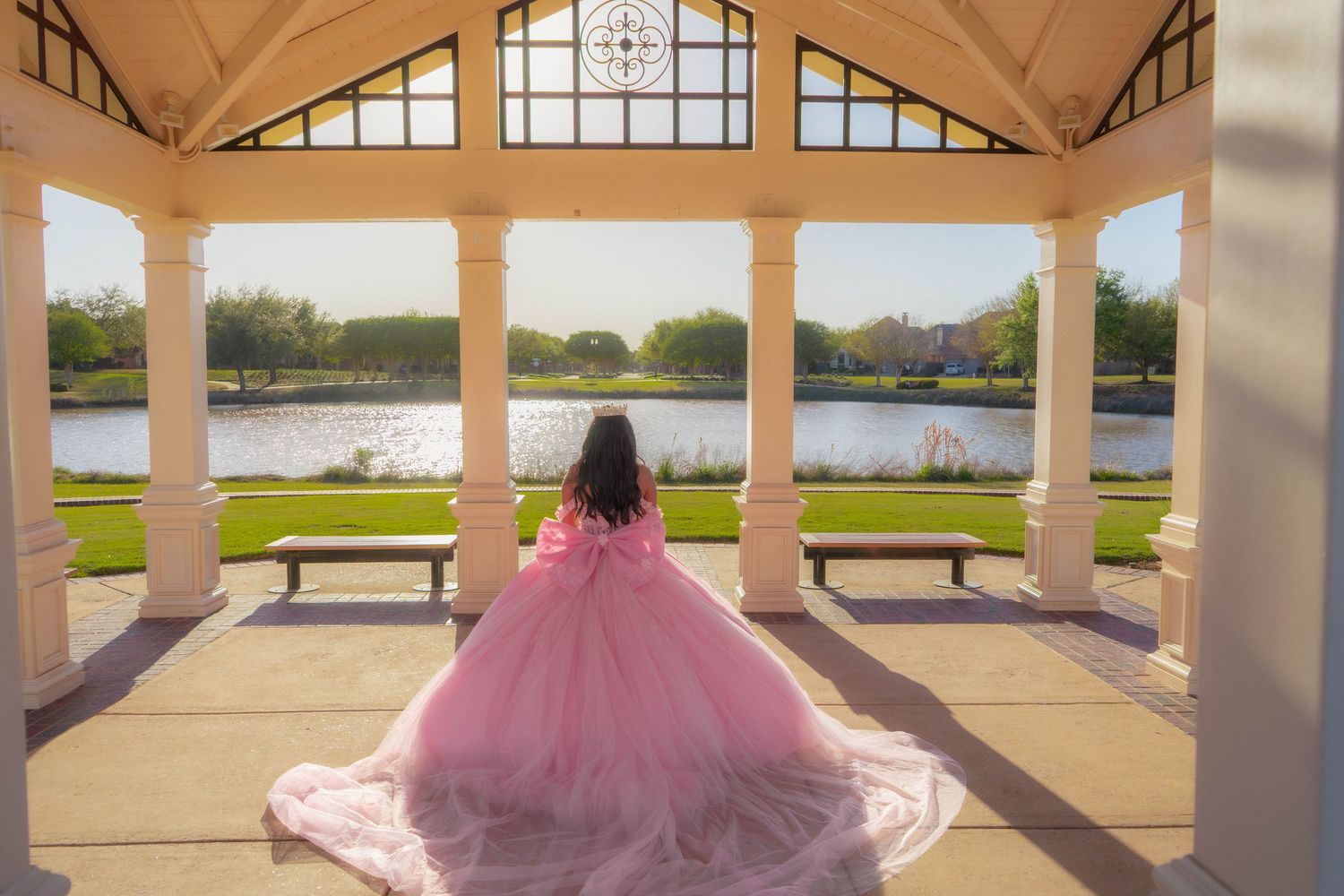 Girl in pink ball gown stands under elegant pavilion overlooking serene lake at golden hour.