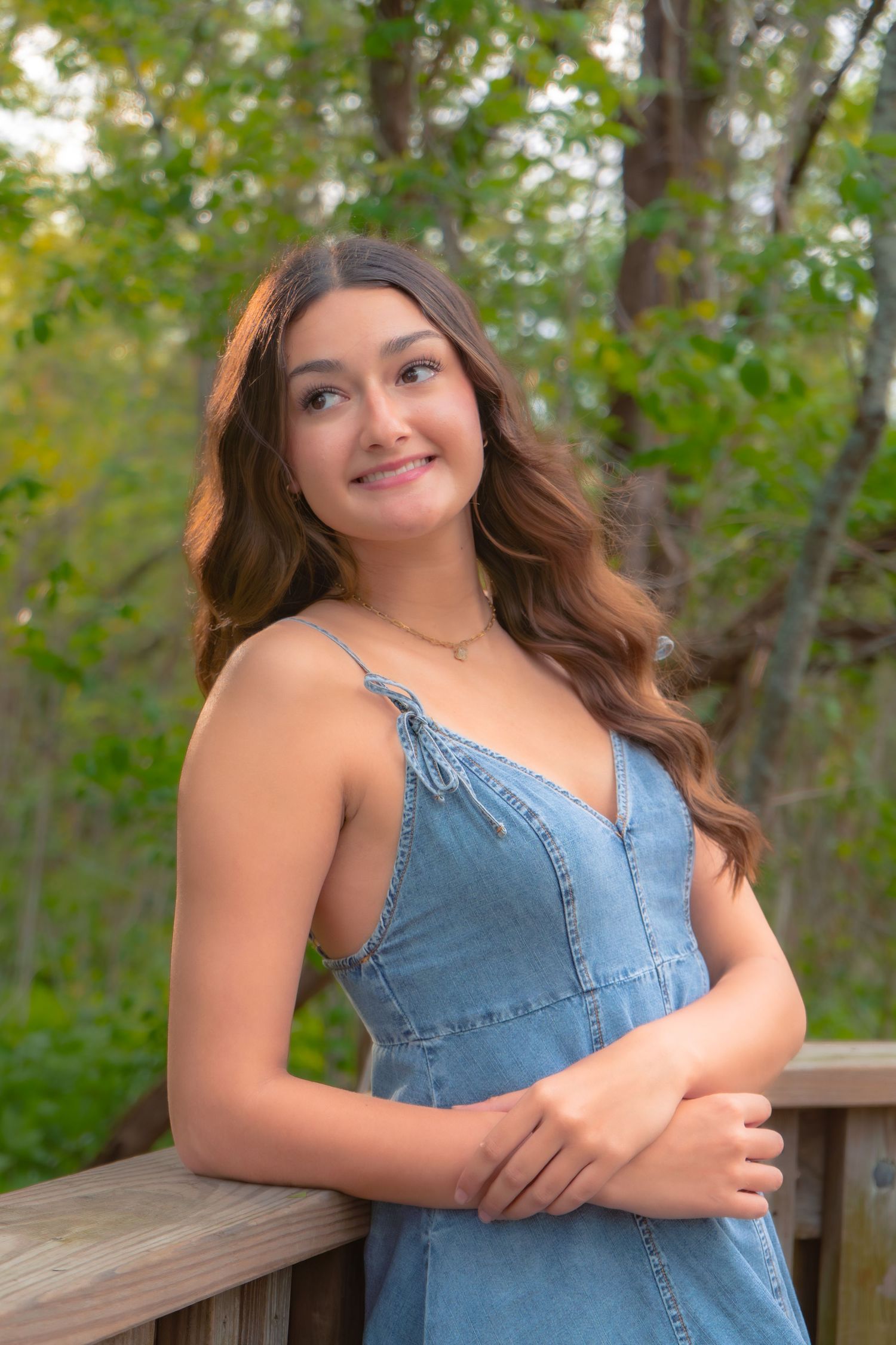 Young woman in denim dress smiling on wooden deck surrounded by lush green trees.