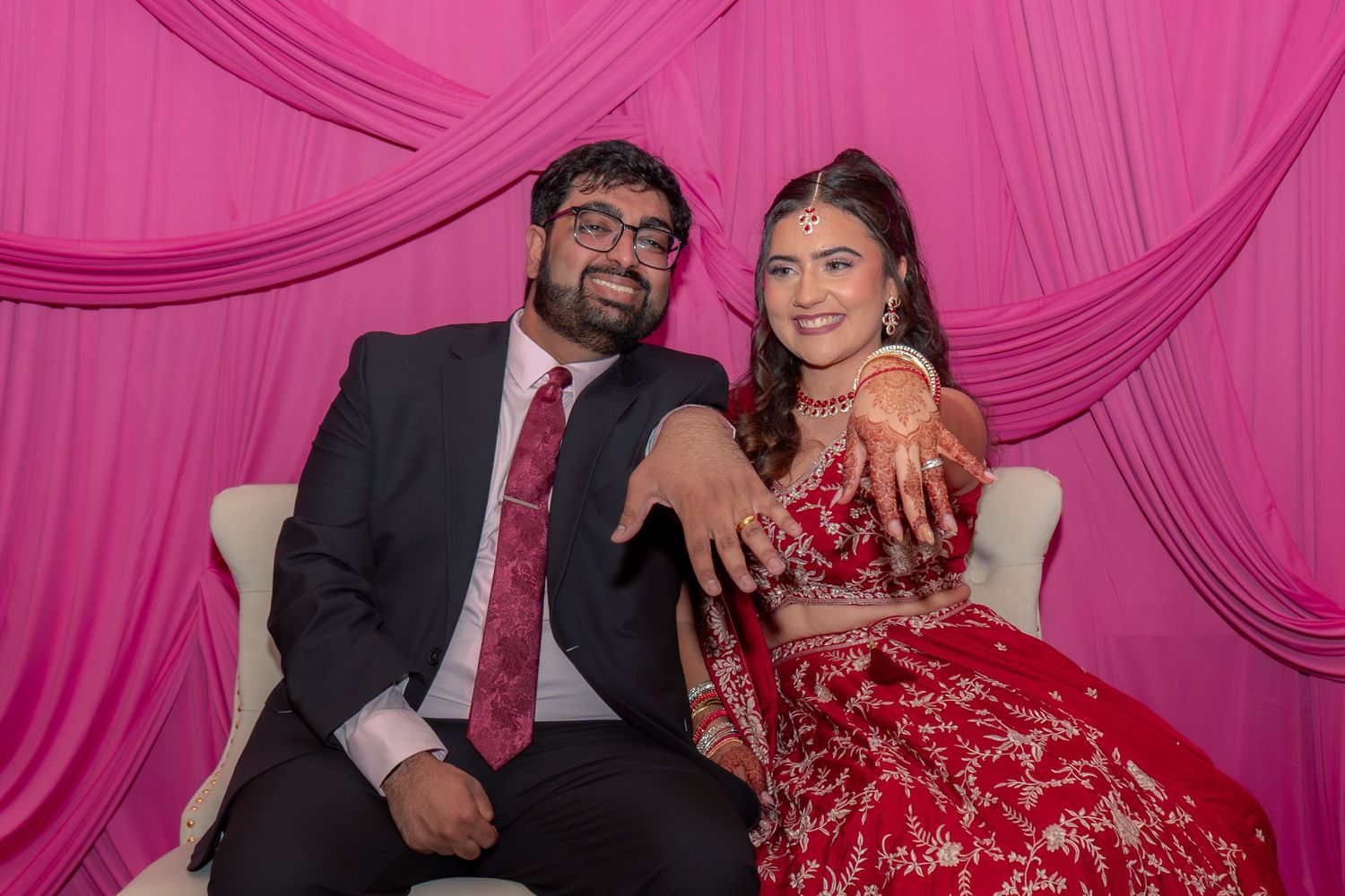 Indian couple in wedding attire posing together, bride in red lehenga with mehndi, groom in dark suit, pink backdrop.