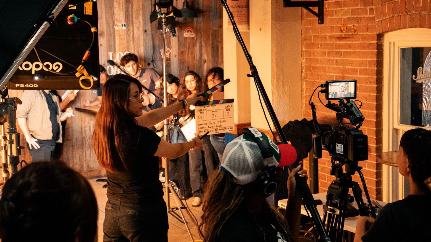 A film director gestures while crew members operate cameras on a brick-walled set.